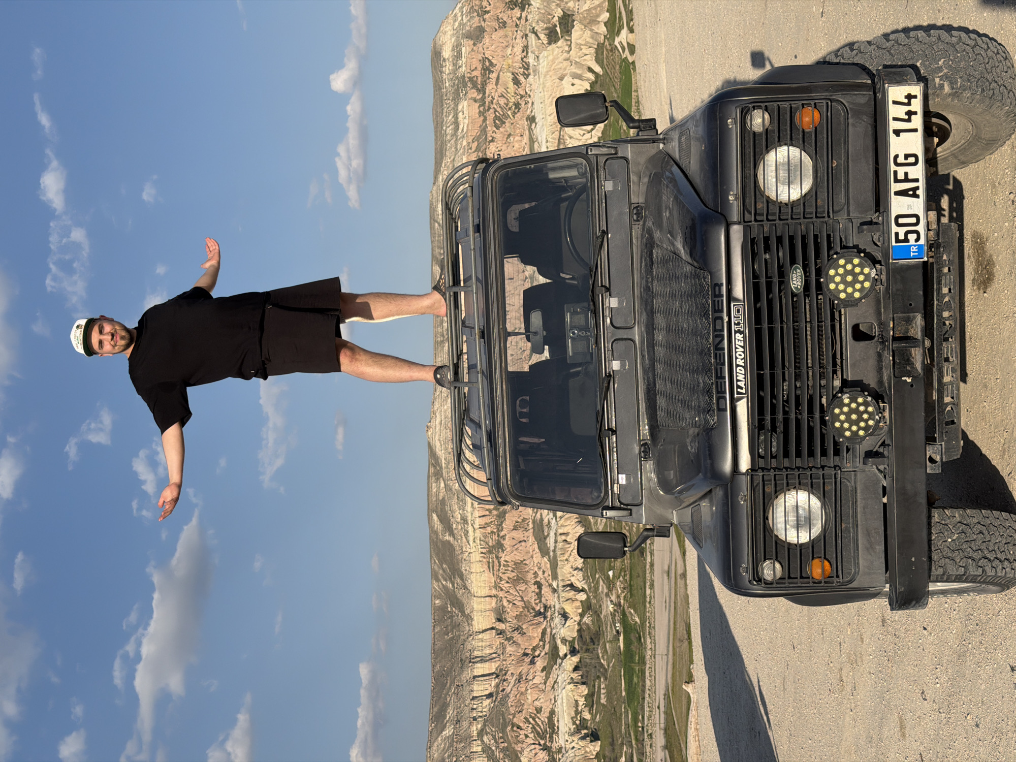 The image, presented as if correctly oriented, shows a man balancing on the roof rack of a dark grey Land Rover Defender 110 in a scenic arid landscape. The man, wearing a black t-shirt, black shorts, and a white baseball cap with an indistinct green logo, stands upright with arms outstretched horizontally, looking towards the camera with a confident or playful expression. His bare legs are visible. The Land Rover Defender 110, an off-road SUV with a robust roof rack, prominent grille, circular headlights, and two circular LED auxiliary lights, is parked on a light-colored road.
In the background, distinctive light-colored rock formations and valleys, typical of Cappadocia, are visible under a bright blue sky with scattered white clouds, suggesting clear, sunny weather during the day. The location is Göreme, Turkey.
Visible text on the vehicle includes 'DEFENDER 110' and 'LAND ROVER' on the grille. The Turkish license plate clearly displays 'TR 50 AFG 144'.
