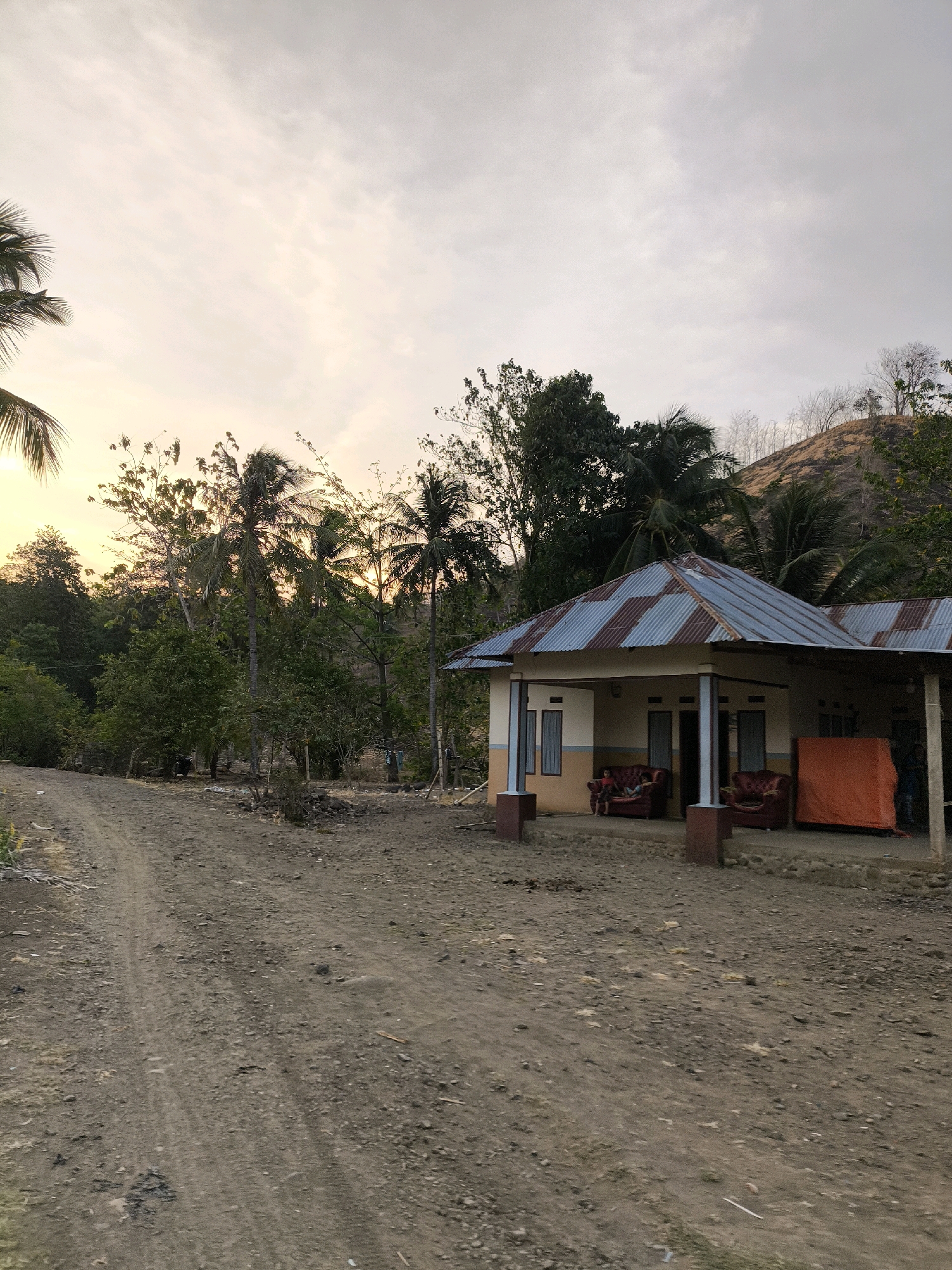 A rural scene at dusk in Unknown, Indonesia, features a simple house with a corrugated metal roof, flanked by two red armchairs and an orange tarp. The house is situated on a slight incline, with a dirt road leading towards it from the left of the frame. The surrounding environment is characterized by lush vegetation, including palm trees and other foliage, against a backdrop of a hilly landscape. The sky is overcast with soft, diffused light, suggesting the sun is setting or has recently set. Two individuals are seated in the armchairs on the porch of the house.
