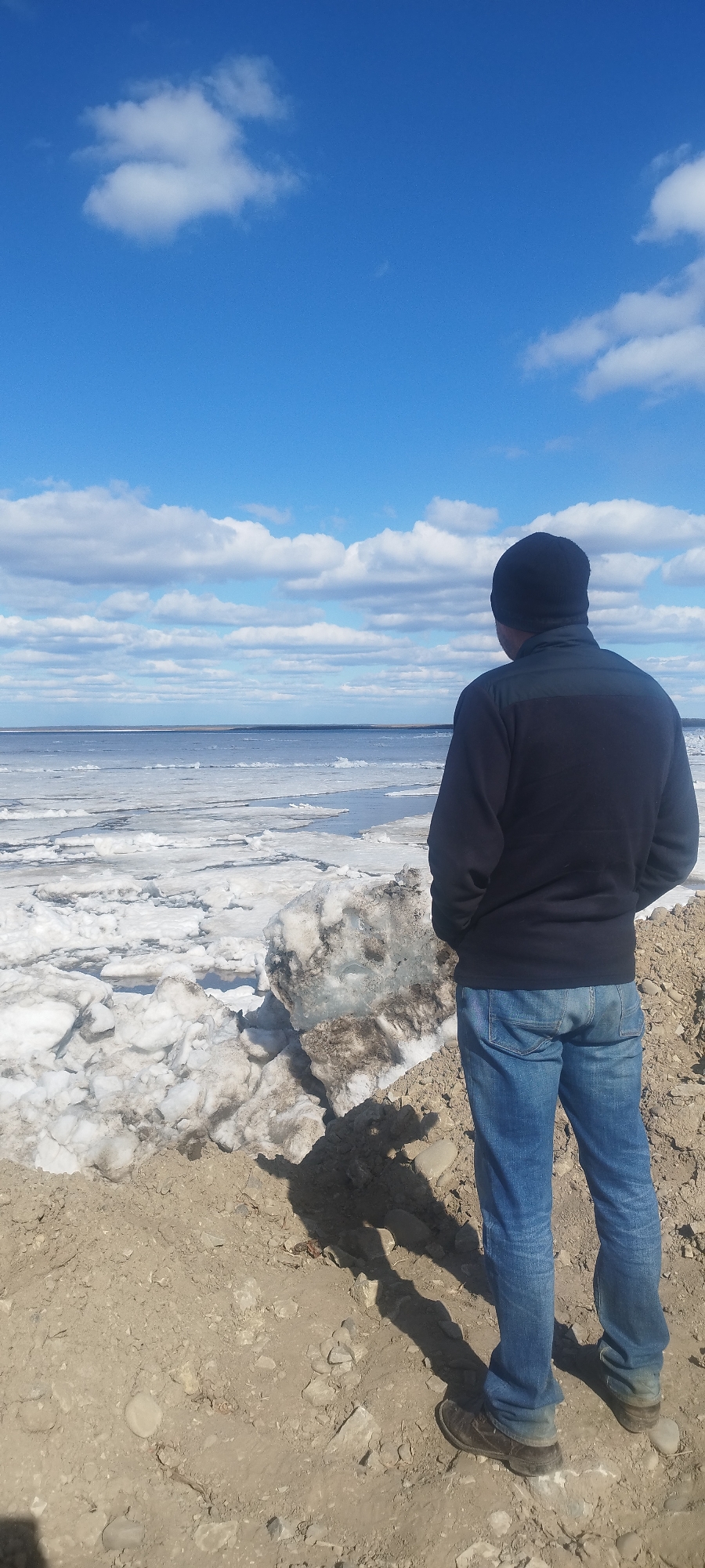 A man stands on a dirt embankment overlooking a body of water covered in ice floes. He is wearing a black beanie, a dark blue jacket, and blue jeans. The sky is bright blue with scattered white clouds. The scene is likely near a river or large lake in a cold climate, potentially near Yakutsk, Russia. There are no visible buildings or other landmarks to confirm the specific location. The man is facing away from the camera, observing the icy landscape.