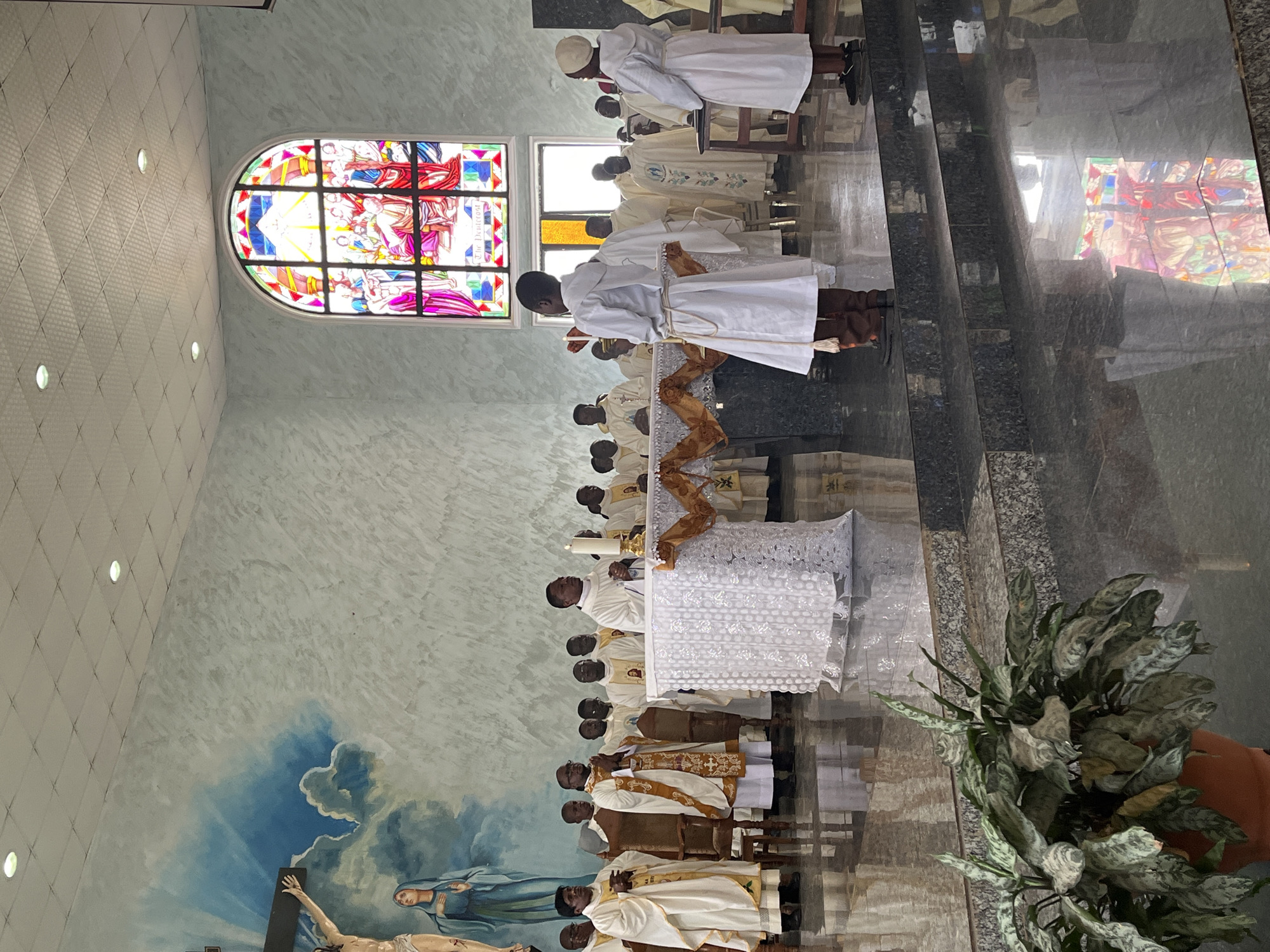 The image shows a religious ceremony taking place inside a church in Vom, Nigeria. A group of men, likely clergy or congregants, are dressed in white robes. Some are standing at an altar, while others are seated or standing in the background. A stained-glass window depicting a religious scene is visible at the back of the church, as is a mural of a crucifixion scene on the left wall. The floor is polished and reflective, showing the lighting and the stained-glass window. A plant is in the foreground. The overall atmosphere appears solemn and reverent.