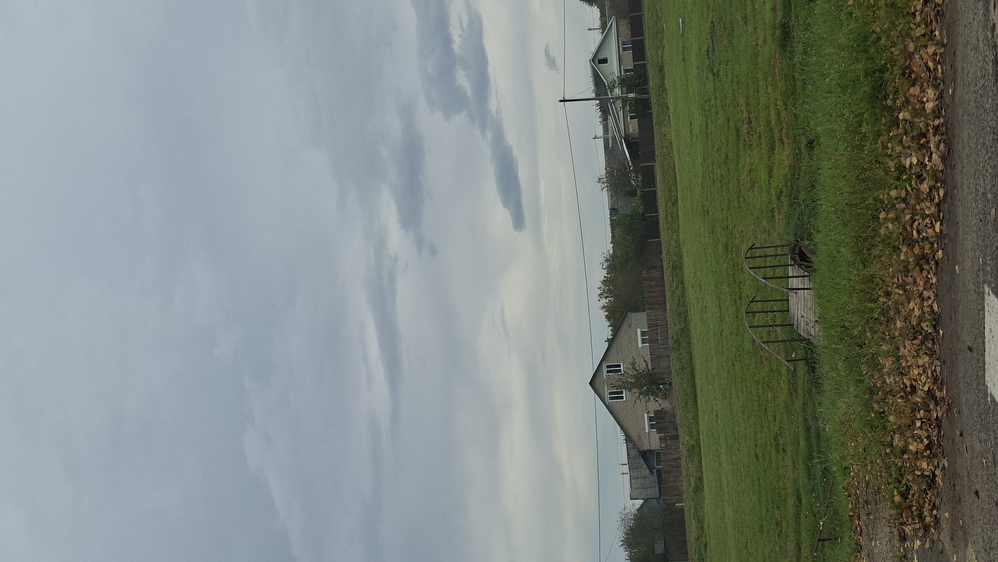 The image depicts a rural scene under a cloudy sky. In the foreground, a small wooden bridge with metal railings spans a grassy ditch. Fallen leaves are scattered on the ground near the bridge. Further back, a green field stretches towards a line of houses and trees. One house, with a gabled roof and two windows, is made of light-colored brick. A wooden fence runs in front of the houses. Power lines are visible above the fence. The overall impression is one of a quiet, overcast day in a village.