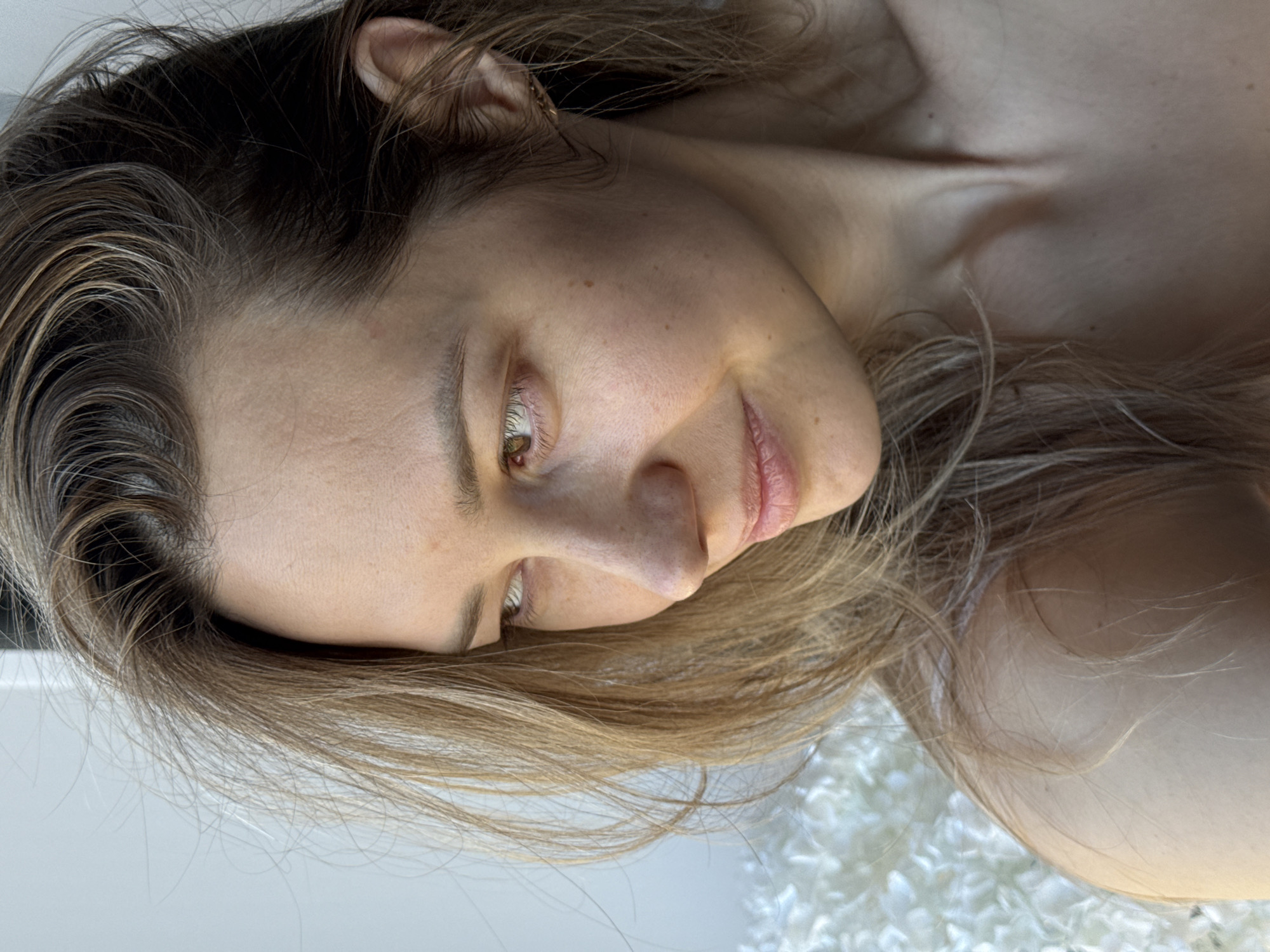 The image shows a woman with light brown hair, fair skin, and brown eyes. She is lying down, looking slightly downward and to the left. Her head is tilted to the left. The background is blurred, showing a white, textured surface. The image provides no contextual information about the city of Chaikovsky, Russia.