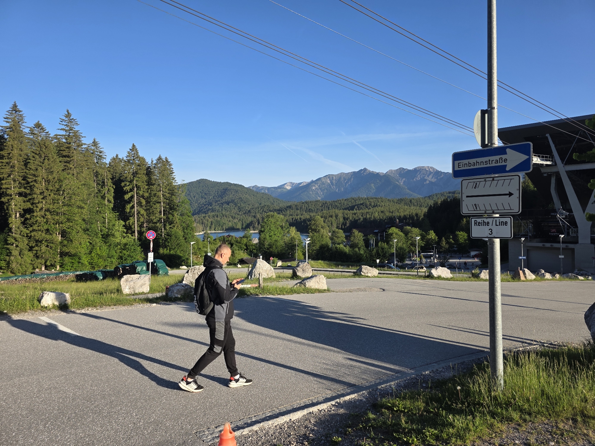 A man, wearing a black and grey hooded jacket, black pants, and black and white sneakers, walks across an asphalt road. He is carrying a black backpack and looking at his phone. The road is bordered by a grassy area with several large rocks. In the background, there is a forest of evergreen trees, a body of water, and mountains under a clear blue sky. A signpost with German text and arrows is visible on the right side of the image, near a building with a modern architectural style. Overhead, cables are strung across the sky.