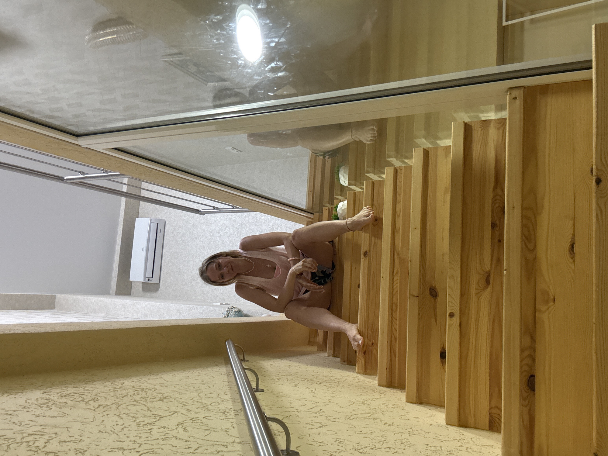 A woman with blonde hair and glasses is sitting on a wooden staircase. She is wearing a pink tank top and dark shorts. She is looking at the camera. The staircase is indoors and appears to be in a modern building. There is a glass wall to the right of the staircase, and a metal railing on the left. An air conditioner is visible on the wall in the background.