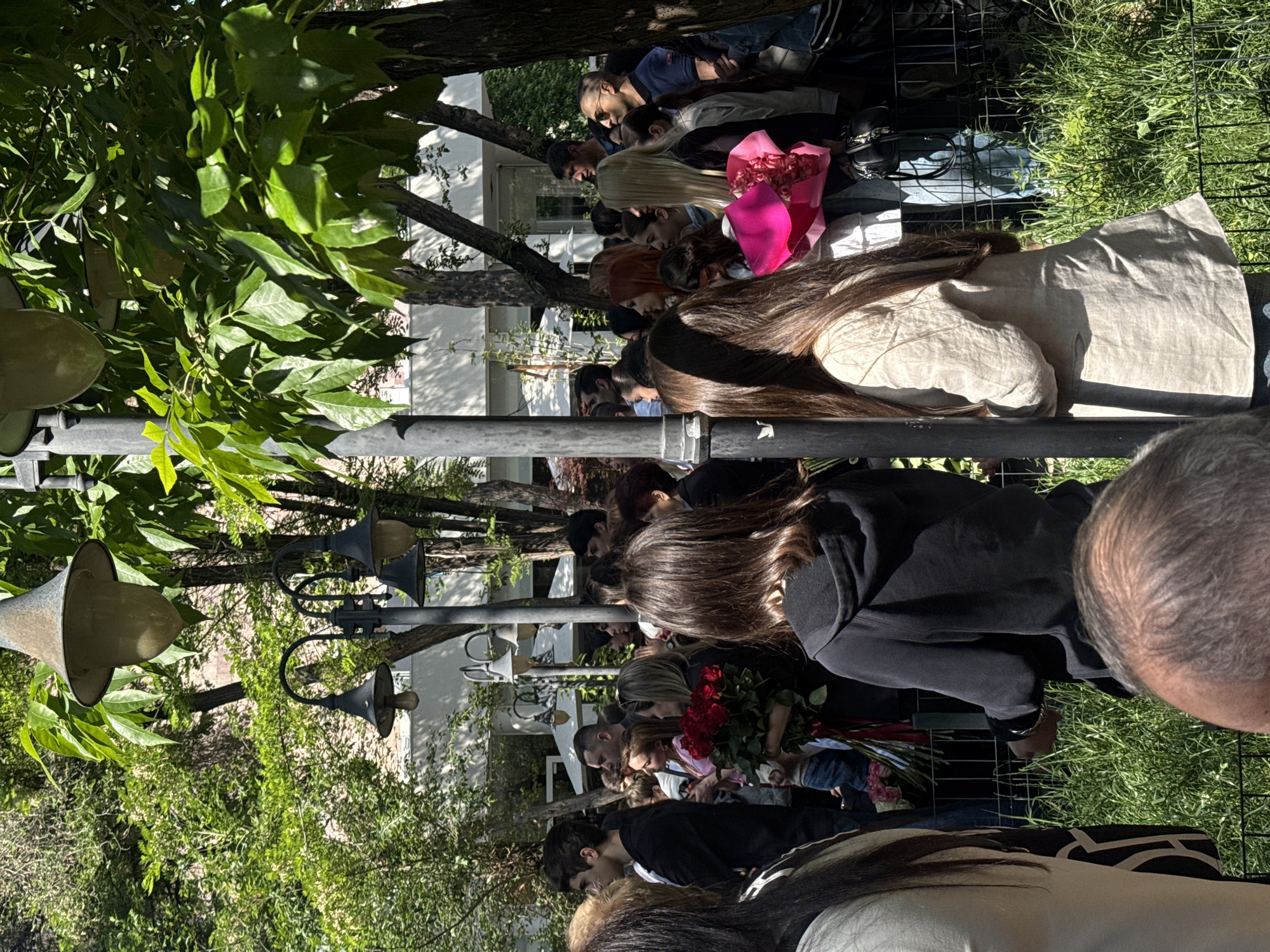 The image shows a line of people in Yerevan, Armenia, waiting behind a black metal fence. Many are holding flowers, including red roses and bouquets wrapped in pink paper. Some people are looking down, and others are obscured by those in front of them. The scene is outdoors, with trees and green foliage visible. Lampposts are interspersed among the trees. White buildings are visible in the background. The atmosphere appears somber and respectful.