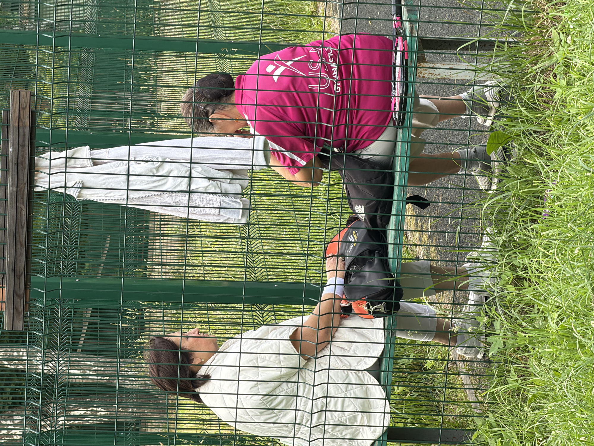 Two women are sitting on a green metal bench in an outdoor setting. The woman on the left is wearing a light-colored quilted jacket and light-colored pants. She has dark hair and is looking to her left. She has a white wristband on her left wrist. Next to her on the bench is a black and orange sports bag.
The woman on the right is wearing a bright pink t-shirt with a white logo and white trim. She has dark hair pulled back in a bun. She is wearing light-colored shorts and grey socks with grey sneakers. She is looking to her left, towards the other woman. A tennis racket with pink strings is visible on the bench to her right.
Behind the bench is a green metal fence. A white jacket is hanging on the fence behind the woman on the left. Trees and foliage are visible in the background, suggesting a park or sports facility. The ground in front of the bench is covered with grass. The scene appears to be in Bykovo, Russia, possibly at a sports complex or park.