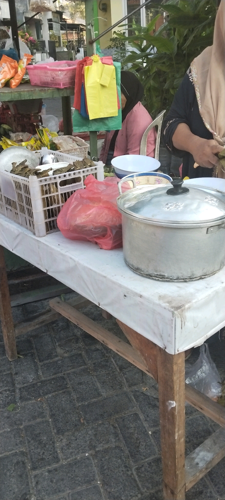 This is a medium shot, outdoor, vertical, eye-level photograph of a street vendor's stall in Surabaya, Indonesia. The stall consists of a white-covered table with wooden legs. On the table, there are several items including a large metal pot with a lid, a red plastic bag filled with food, a white plastic crate containing food items wrapped in brown leaves, a white bowl, and a light blue bowl. In the background, there are colorful plastic bags hanging on a green pole, and a person wearing a pink hijab and a pink shirt is seated. To the right, another person wearing a light brown hijab and a dark blue garment is standing and appears to be preparing food. The ground is paved with dark grey rectangular bricks. The lighting suggests it is daytime, possibly late afternoon, with the sun casting some shadows.