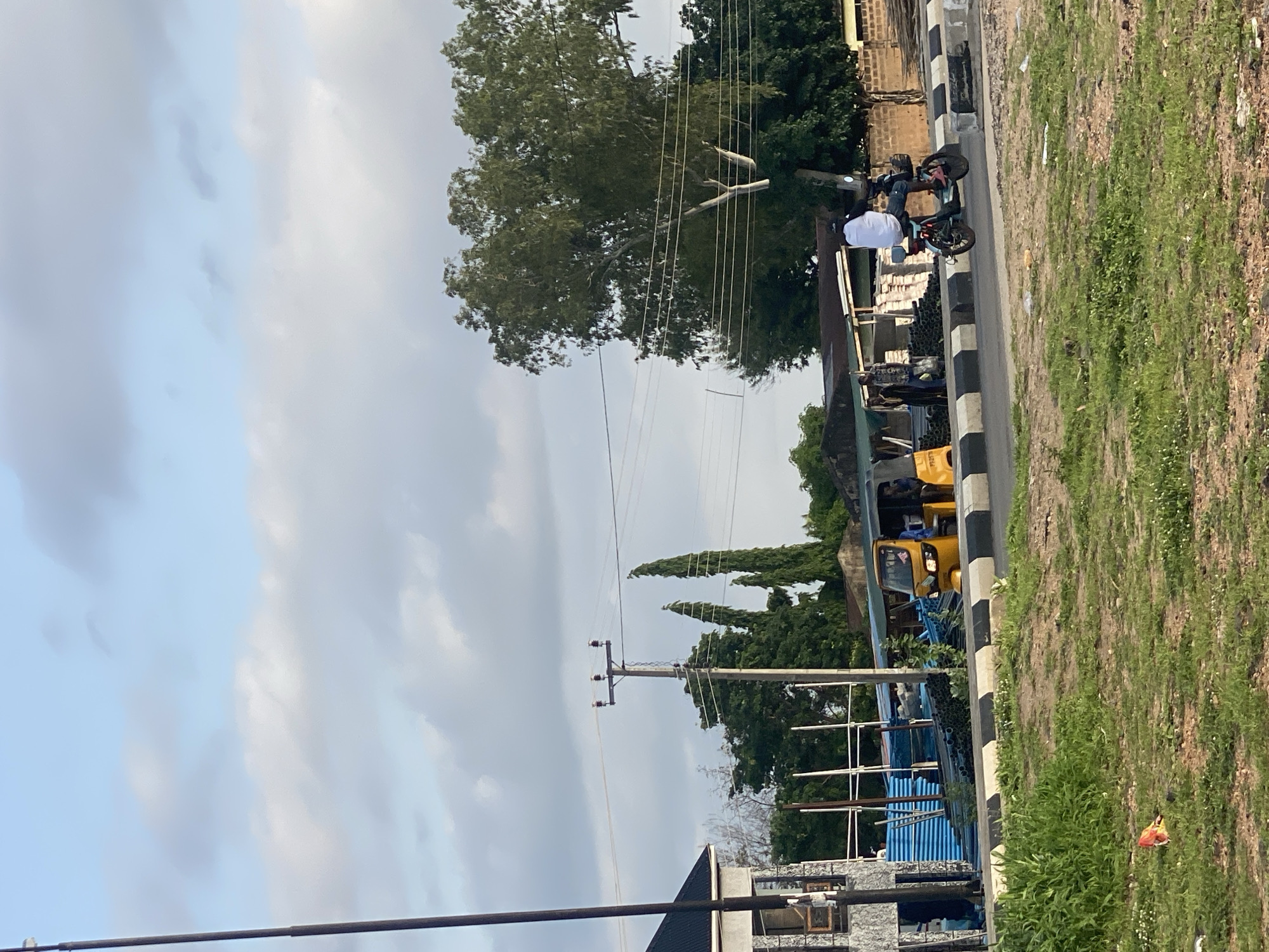This image captures a street scene in Jalingo, Nigeria, under a partly cloudy sky. On the right side of the road, a person is riding a motorcycle. They are wearing a white shirt and dark shorts, with their back to the camera. The motorcycle is dark in color.
To the left of the motorcycle, a yellow auto-rickshaw, also known as a "Keke Napep," is parked at the side of the road. It appears to be stationary, with at least one person visible inside the driver's cabin. Several other individuals are standing near the auto-rickshaw, some under a makeshift shelter or awning.
In the background, there are various structures, including buildings with corrugated iron roofs and a brick wall. Piles of what appear to be construction materials, possibly pipes and metal sheeting, are stacked along the roadside. Electrical poles and overhead wires are visible, indicating the presence of utilities.
The foreground consists of a grassy area with some patches of bare earth, suggesting a natural or undeveloped space adjacent to the urban infrastructure. A piece of red and white litter is visible on the grass in the lower left portion of the frame. The road itself is paved and features a distinctive black and white striped curb. The overall impression is a typical street view in a Nigerian city, showcasing local transportation and commercial activity.