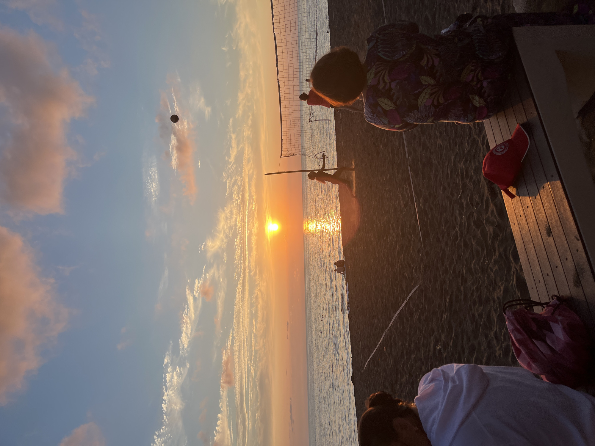 The image depicts a beach volleyball game at sunset on Ureki beach, Georgia. The sun is low on the horizon, casting a warm orange glow over the sea and the sky, which is adorned with wispy clouds. A volleyball net is set up on the dark sand, and several people are engaged in playing. One player is mid-air, about to hit the ball, while another is positioned behind the net. In the foreground, two individuals are seated on a wooden bench, observing the game. One person, viewed from behind, wears a colorful patterned top, and next to them, a red cap rests on the bench. Further in the background, near the water's edge, a few other people are visible, some possibly swimming or wading. The scene conveys a sense of leisure and twilight enjoyment on a coastal setting.