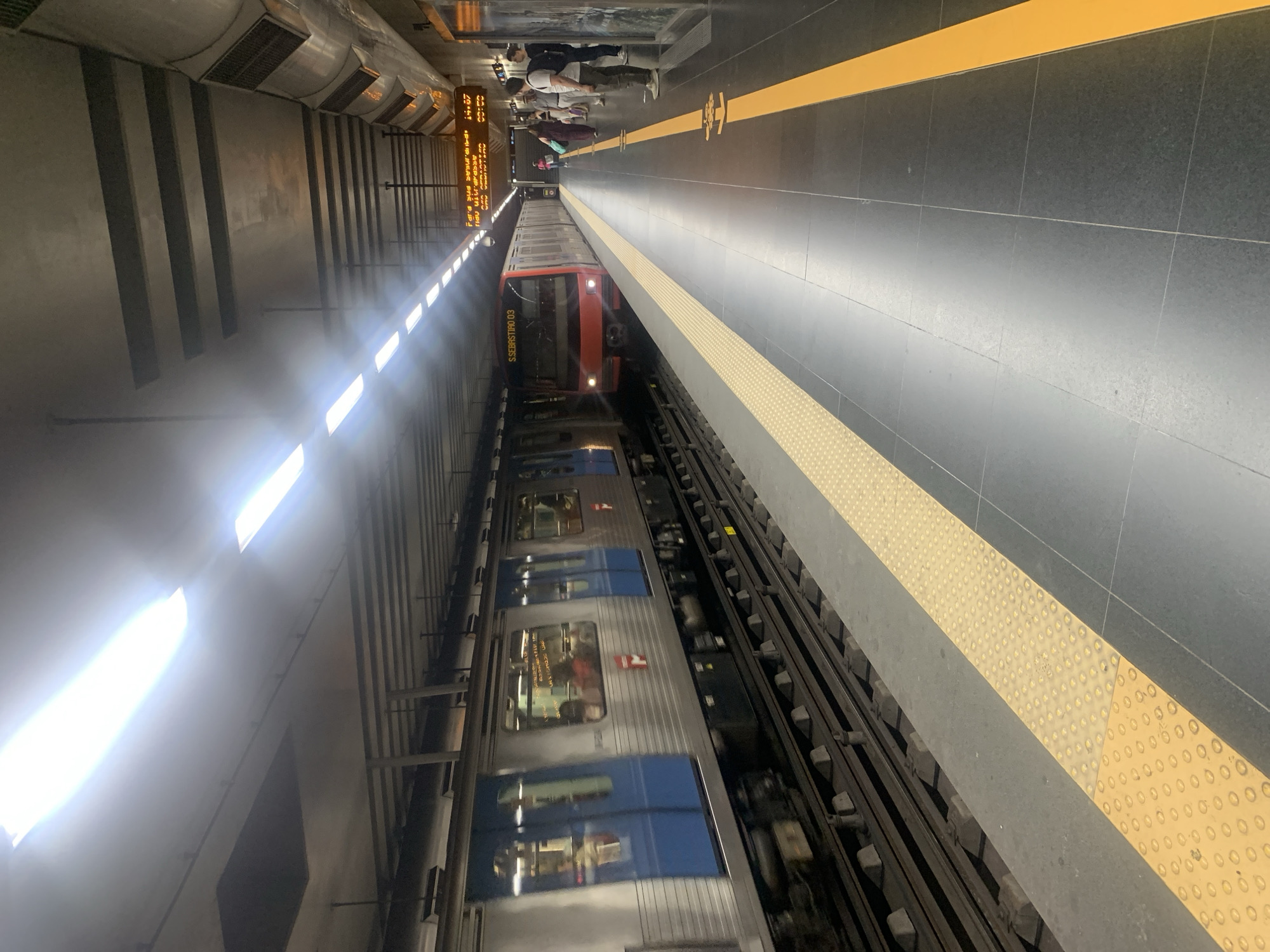 The image captures a view of a subway station in Lisbon, Portugal. A subway train with a red front and silver carriages is stopped at the platform. The train has "S. SEBASTIAO 03" written on its front. Passengers are visible through the train windows. A yellow line marks the edge of the platform, and a tactile paving surface is present. In the background, people are waiting on the platform, and a digital display board shows train information. The station appears to be underground, with bright fluorescent lighting illuminating the scene. The atmosphere seems ordinary, with commuters waiting for their trains. The time of day is not explicitly stated, but the lighting suggests it could be daytime or well-lit indoors. The overall scene depicts a typical urban subway environment.