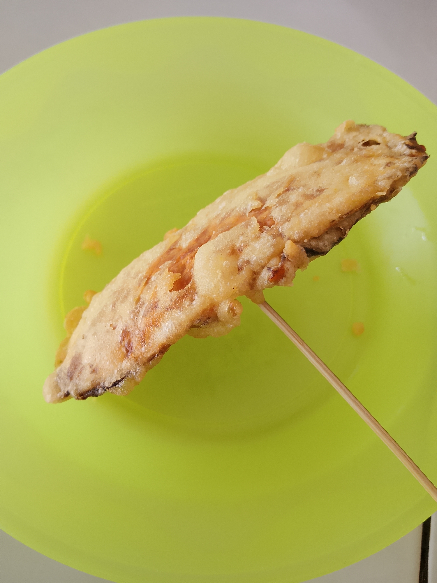 A single piece of fried banana is skewered on a thin wooden stick, resting diagonally on a bright green plate. The banana is coated in a golden-brown batter and appears slightly translucent in places, revealing the fruit underneath. Small, crumbly bits of batter are scattered on the plate around the banana. The background is a plain, light gray surface, likely a table or countertop. The scene is indoors, suggesting a domestic setting or a small eatery, possibly in Balun, Indonesia, given the mention of the city. No people or other objects are visible in the image. The lighting is even and appears to be natural or from an overhead source.