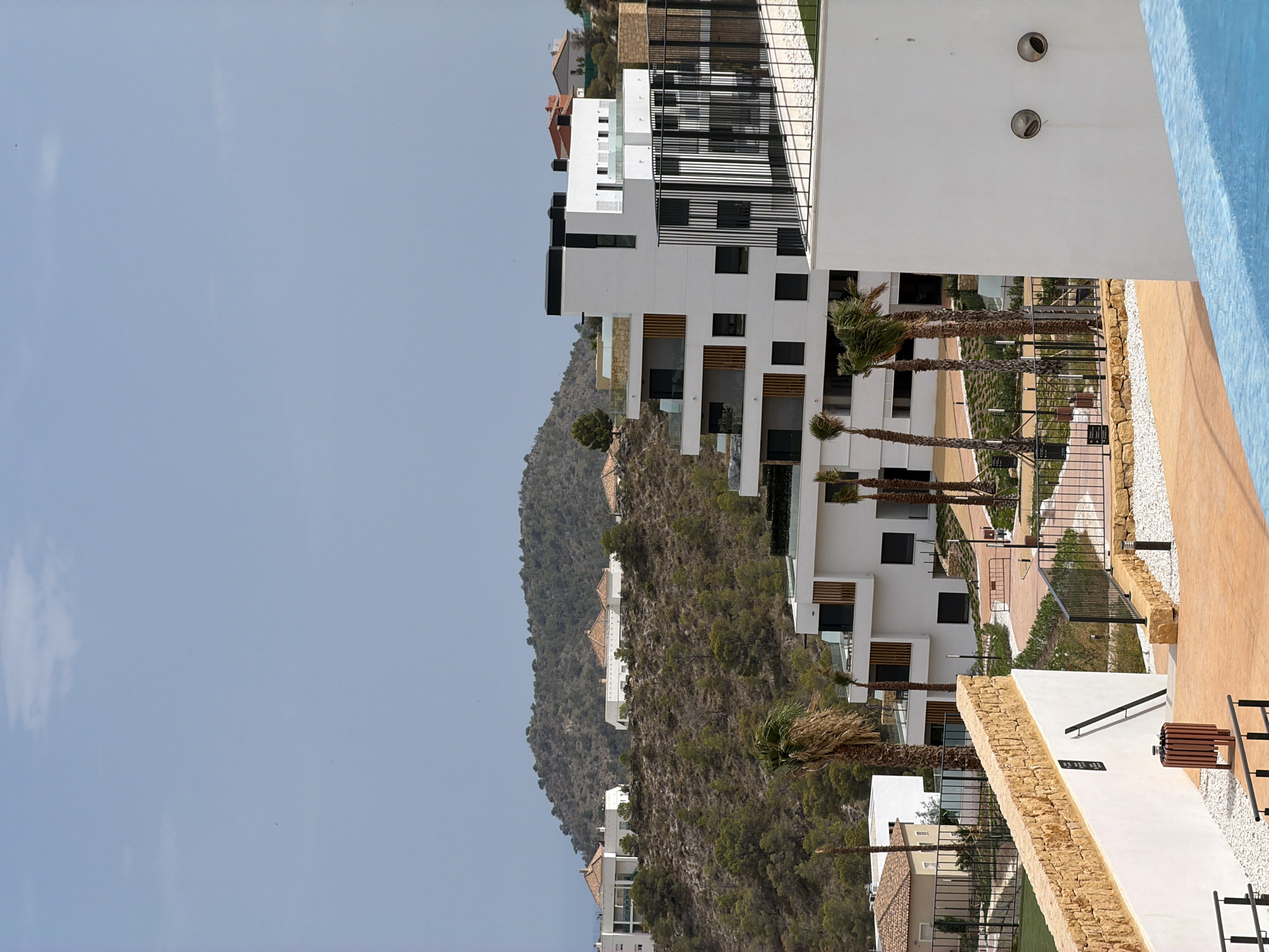 The image depicts a modern residential complex nestled in the hilly terrain of Finestrat, Spain. In the foreground, a bright blue swimming pool edge is visible, suggesting a leisure area. To the right, a white building wall with two circular vents is prominent. A pathway of orange concrete and white gravel leads towards the buildings.
Further back, a terraced white apartment building with multiple balconies and dark-framed windows dominates the midground. Palm trees are scattered throughout the complex, adding to the Mediterranean ambiance. The buildings are situated on a hillside covered with vegetation and rocky outcrops.
In the background, more white residential buildings with terracotta roofs are scattered across the lower slopes of a larger, forested hill. The sky is a pale blue with a few wispy clouds. The scene conveys a sense of contemporary development in a natural, elevated landscape characteristic of the Finestrat region, known for its coastal proximity and scenic views. There are no people or animals visible in the image.