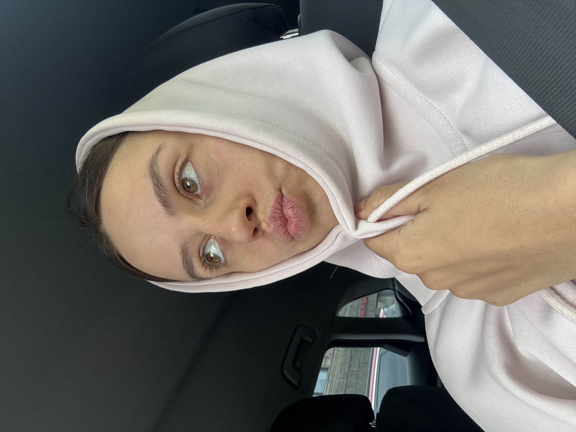 A woman with brown hair and eyes wearing a light pink hoodie is in the passenger seat of a car. She is wearing a seatbelt. The background visible through the car window suggests an urban setting, possibly Krasnoyarsk, Russia, as indicated. The woman is puckering her lips and looking at the camera.