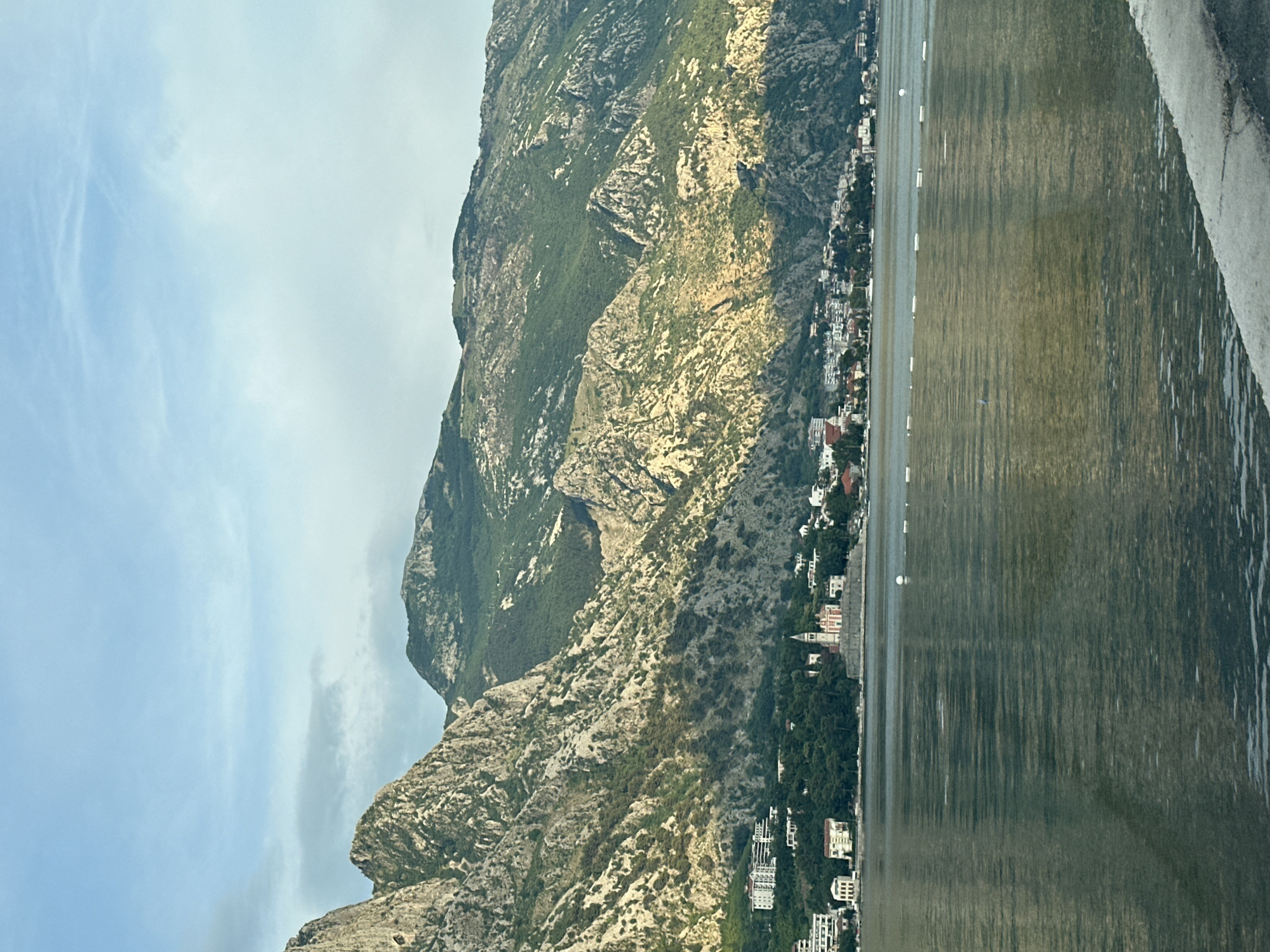 The image captures a view of Prčanj, Montenegro, with a focus on its waterfront and the surrounding mountainous landscape. The foreground is dominated by the calm, rippling surface of the water, which reflects the light and the colors of the sky. In the mid-ground, a line of white buoys marks a boundary in the water. Beyond the water, the town of Prčanj is visible, characterized by its clustered buildings, including what appears to be a church with a distinct red roof and a tall spire. Dense green trees surround the town, creating a lush contrast with the stone buildings. The backdrop of the scene is formed by the imposing, rugged mountains that rise steeply behind the town. These mountains are a mix of exposed rock faces, showing shades of grey and brown, and areas covered in green vegetation, particularly on the lower slopes. The sunlight illuminates parts of the mountains, creating a play of light and shadow that accentuates their texture and contours. The sky above is a pale blue, with wispy clouds suggesting a clear, perhaps late afternoon or early morning atmosphere. The overall impression is one of a serene coastal settlement nestled at the foot of a dramatic natural landscape.