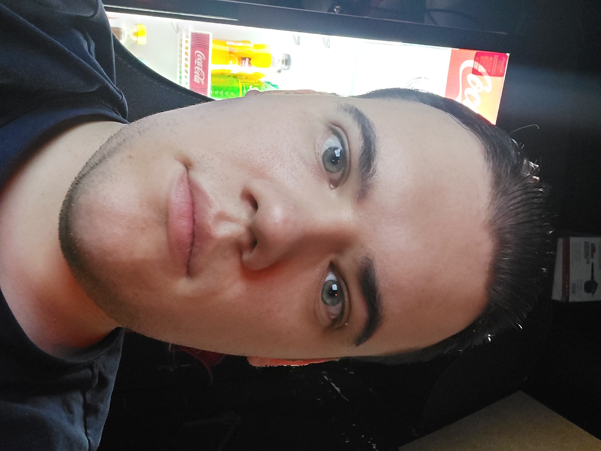 A close-up, low-angle selfie of a light-skinned male with short, dark hair. His eyes are gray-green. He has a short, dark beard. Behind him is a refrigerated beverage display, featuring Coca-Cola branding. The location is Tashkent, Uzbekistan.