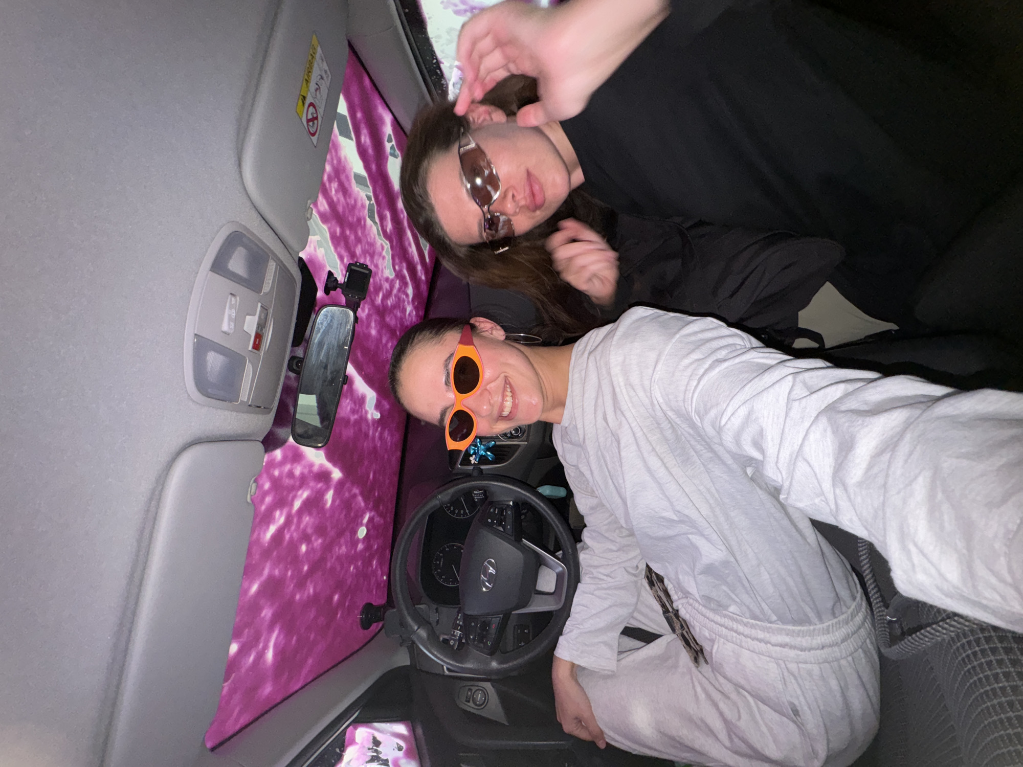Two young adults are in a car wash. One person wears black clothing and brown sunglasses. The other wears white clothing and orange sunglasses. The car's interior includes a rearview mirror, steering wheel, and sun visor. The carwash features pink and white foam. This image was not taken in the city of Krasnoyarsk, Russia.