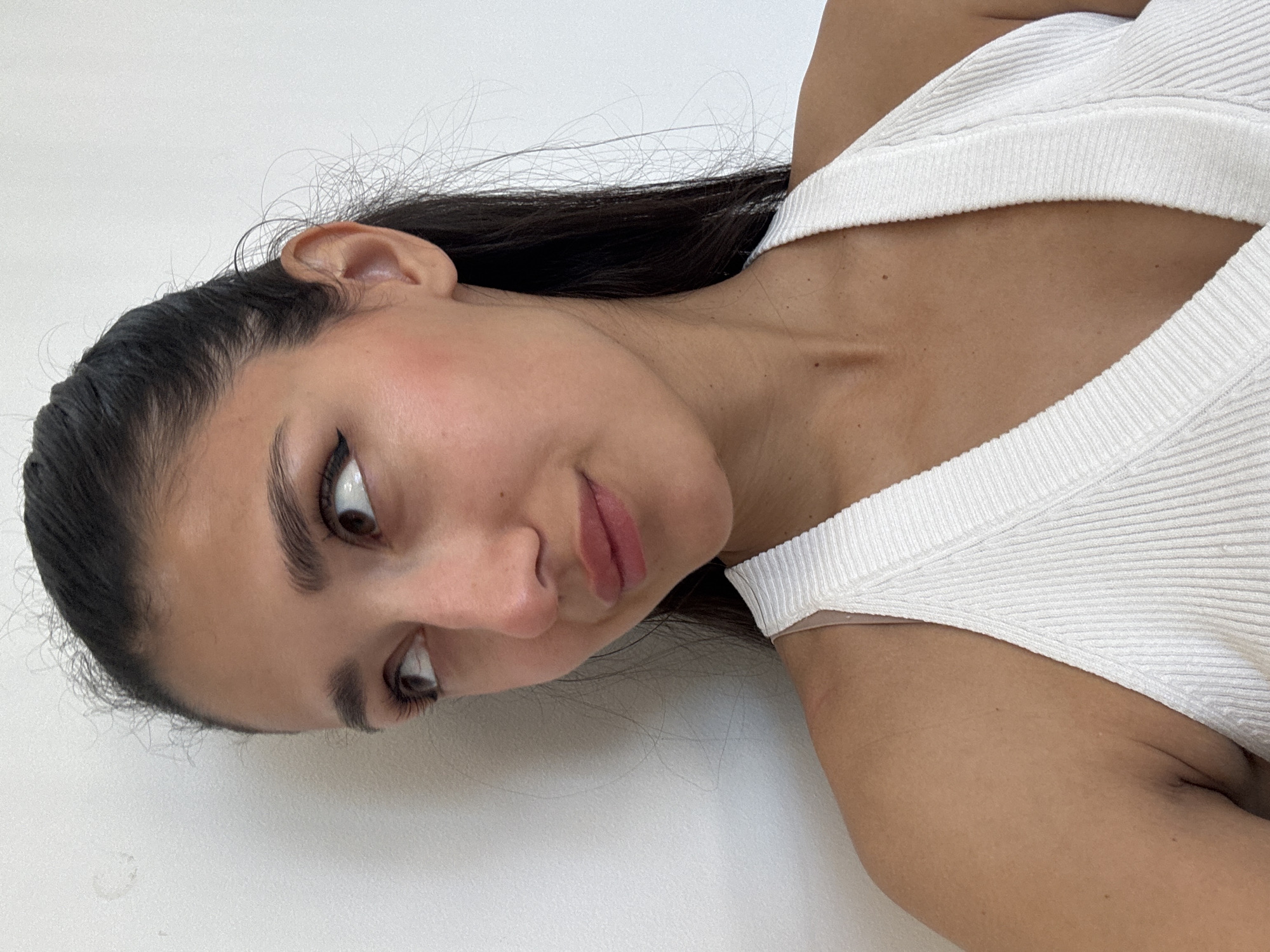 The image features a young adult female with dark, long hair pulled back from her face, with some loose strands around her hairline. Her head is turned to the left, and her gaze is directed upwards and to the left, conveying a thoughtful or subtly amused expression with a slight, closed-mouth smile. She has olive-toned skin and is wearing a white, ribbed V-neck halter top. A thin, light-colored strap, likely from an undergarment, is visible on her right shoulder. Her makeup includes defined eyebrows, winged eyeliner, mascara, blush on her cheeks, and a subtle lip tint. The background is a plain, off-white wall, with a faint, circular, light-brown stain or smudge visible on the upper left portion of the wall behind her head. The scene is illuminated by even, soft lighting. The specific geographical location, including the city of Budva, Montenegro, cannot be determined from the visual information present in the image.