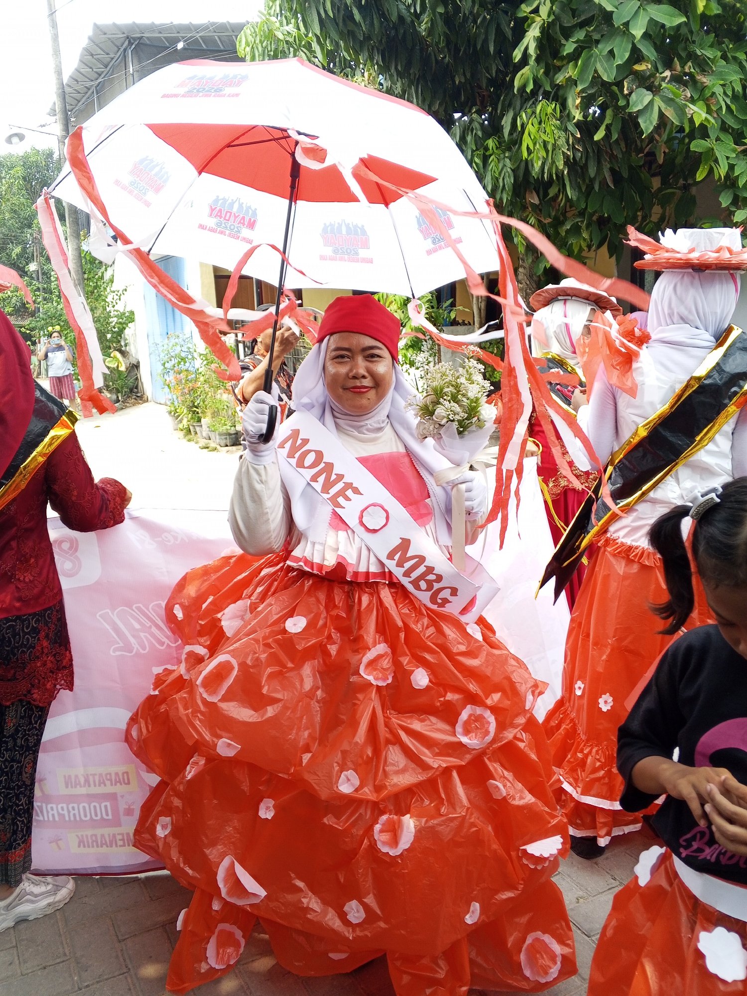 In Ciruas, Indonesia, a woman is participating in a parade, holding an umbrella adorned with red and white streamers. She wears a red headscarf, white gloves, and a large, voluminous orange skirt decorated with white flower-like appliqués. A sash across her chest reads "NONE MBG". She is standing on a paved surface, possibly a street, with other participants in similar colorful attire visible in the background, including a person in a red garment and another wearing a white outfit with an orange ruff. Trees and buildings are visible behind them.
