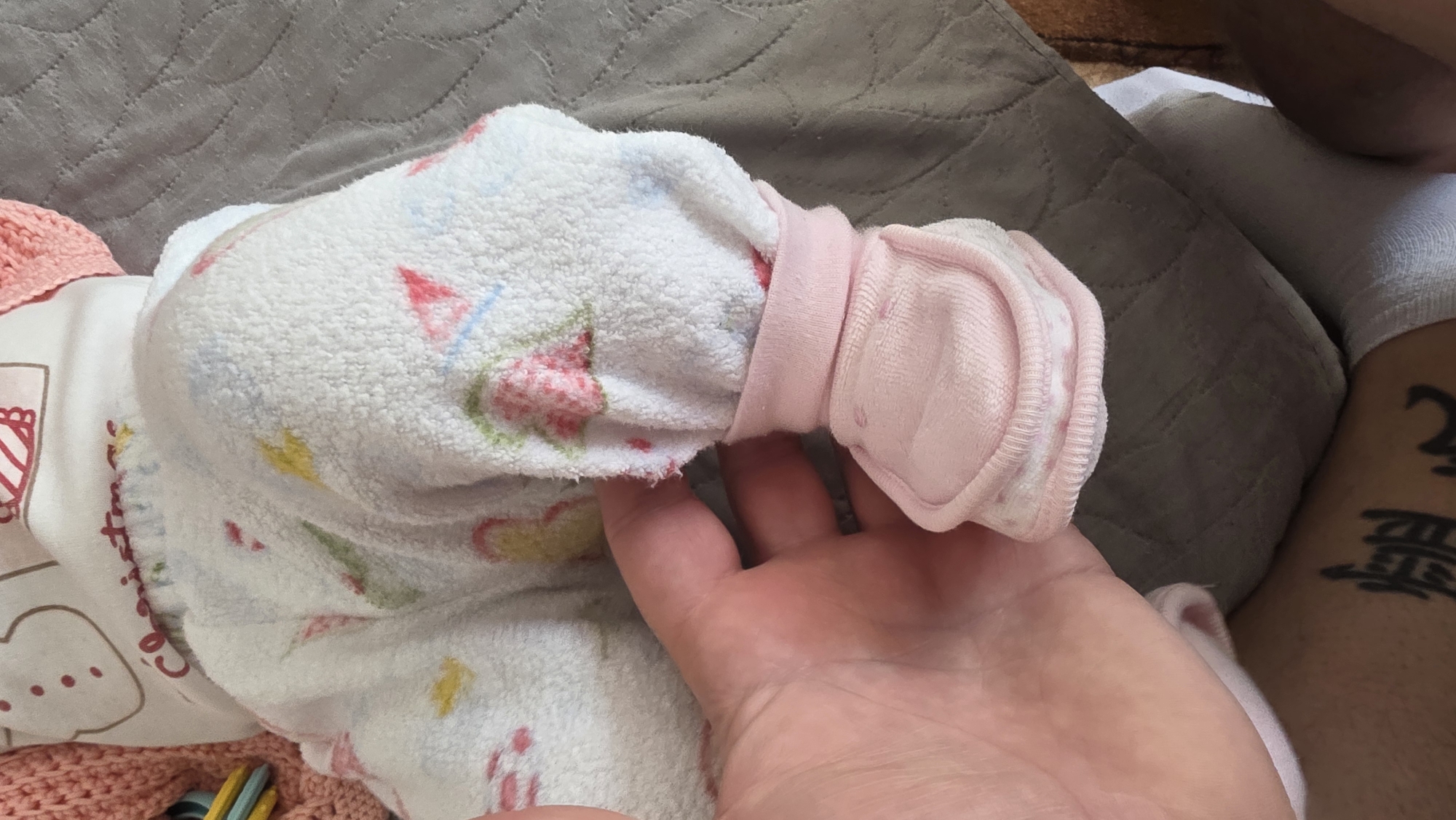 A baby in a white, patterned sleeper with attached mittens is being held. A hand is visible, seemingly adjusting the sleeve or mitten. A portion of a leg with a tattoo is also visible in the background, along with a gray padded surface. There is no information to support the location being Blagoevgrad, Bulgaria.