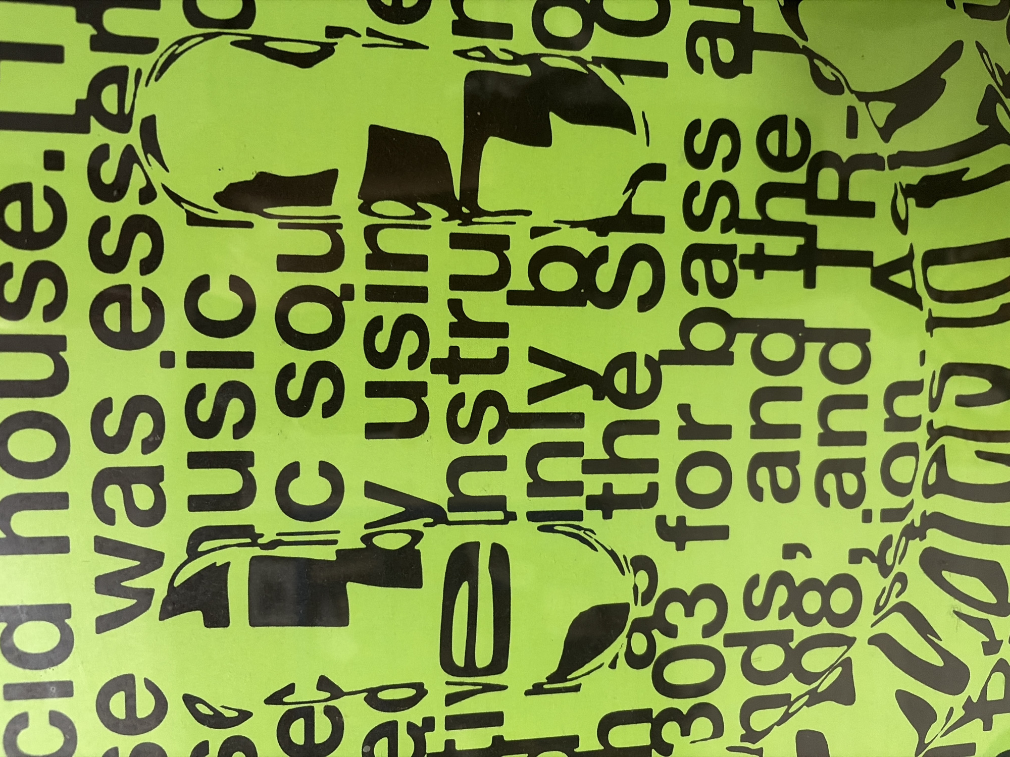The image shows a close-up of a bright green poster with black text and abstract black graphic elements. The text is in all caps and appears to be a list of terms related to electronic music and synthesizers, such as "acid house," "music," "instruments," "303," "909," and "TR-." The graphic elements seem to be stylized depictions of human faces or heads, with some abstract lines and shapes intermingled with the text. The lighting is even, and the focus is sharp, suggesting a well-lit indoor environment. There are no people visible in the image. The poster is mounted on a flat surface, possibly a wall, but the surrounding environment is not discernible. There is no indication of the city of Mataró, Spain, within the image.
