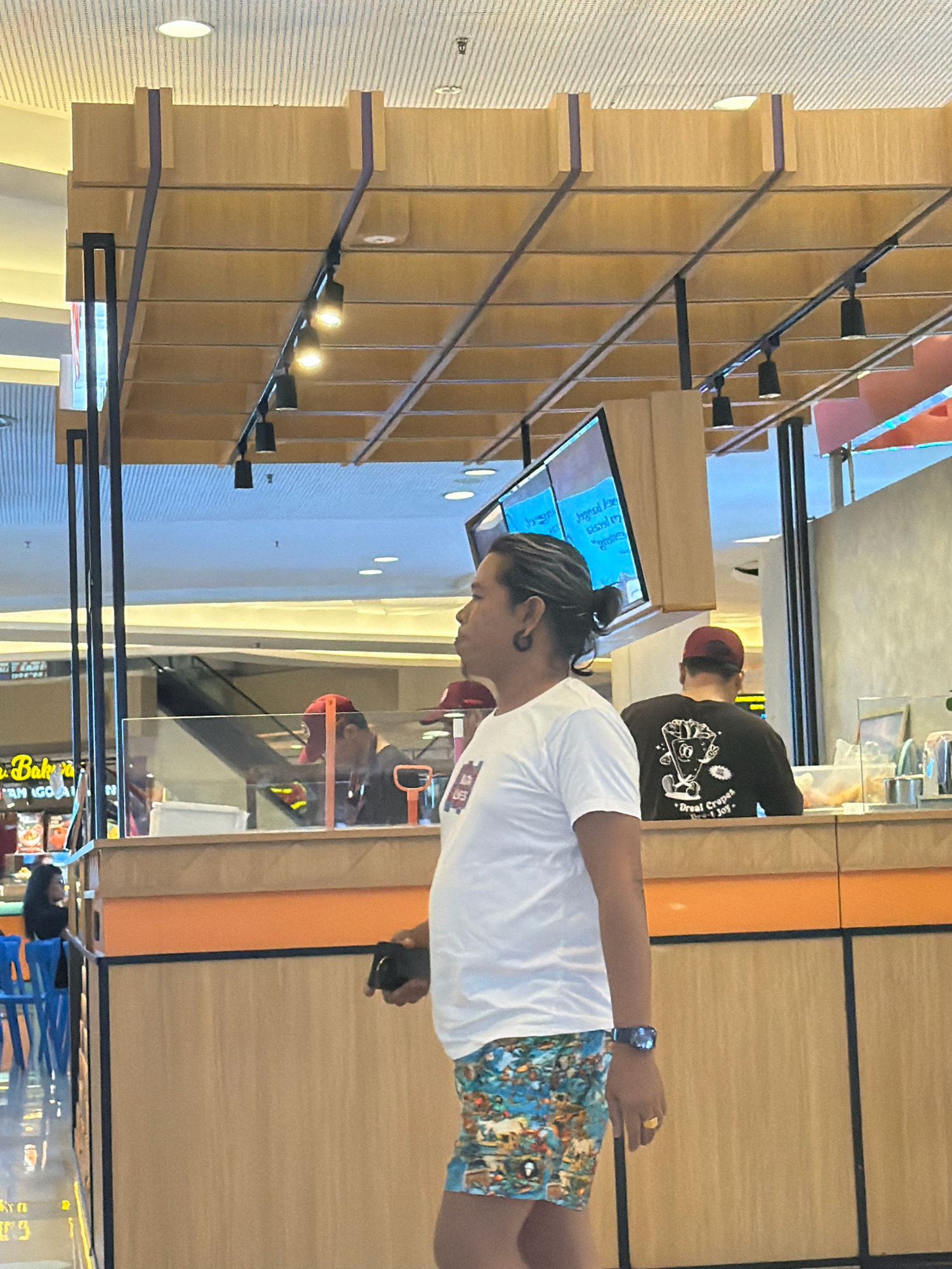 A person stands in the foreground facing right, wearing a white t-shirt, colorful patterned shorts, a watch, and a ring. Their hair is tied back in a bun. In the middle ground, another person wearing a black t-shirt with a graphic print and a red baseball cap is visible from the back. They appear to be at a counter or serving area. To the left, a person in a red hat is partially visible. The environment appears to be an indoor marketplace or food court with wooden paneling, a counter, and an escalator in the background. The lighting is artificial.