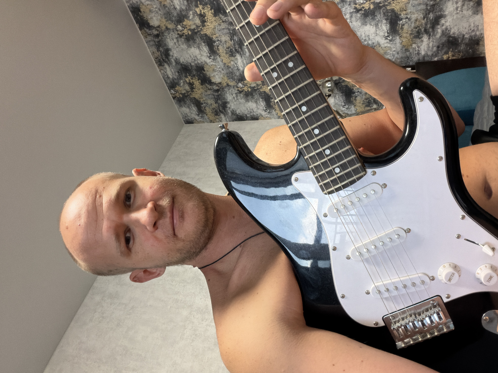 A shirtless, bald, light-skinned man with a beard is holding a black and white electric guitar and looking directly at the camera. He is indoors, in what appears to be a bedroom. The background consists of a textured wall with a dark, abstract pattern and a lighter grey wall. The man's right hand is on the fretboard of the guitar, and his left arm is resting on the guitar body. The lighting suggests natural light coming from the left side, casting a shadow on his shoulder and arm. The scene is consistent with a personal space in Yekaterinburg, Russia, possibly a home or apartment.