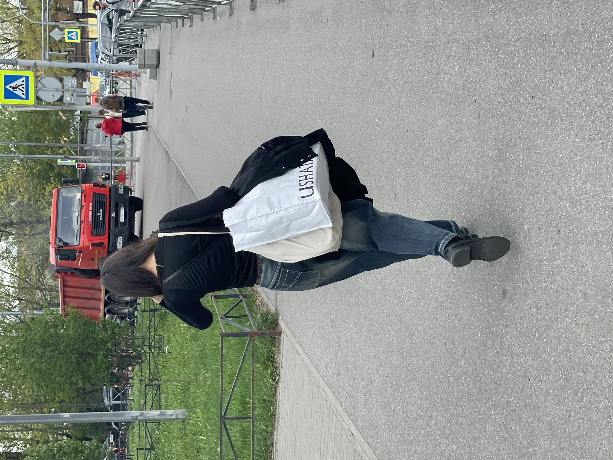 A woman is walking on a sidewalk in Saint Petersburg, Russia. She is wearing a black long-sleeved shirt, blue jeans, and black shoes. She has a white shopping bag over her shoulder. A red truck is parked on the side of the road. There is a pedestrian crossing sign nearby.