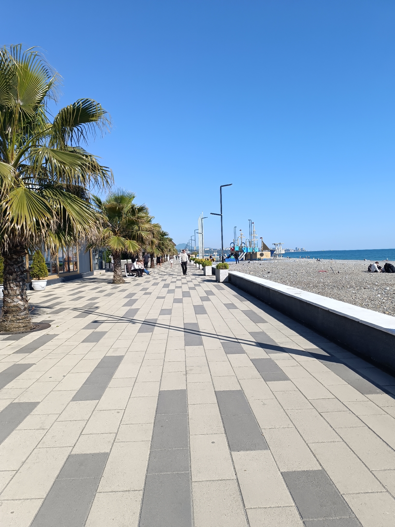 The image shows a wide, paved promenade lined with palm trees on one side and a pebble beach on the other. The promenade is made of alternating rows of light and dark gray rectangular paving stones. On the left side of the promenade are palm trees planted in the ground and some potted plants near a low building. People are visible along the promenade, including individuals walking and sitting on benches. On the right, a low wall separates the promenade from a pebble beach. Several people are sitting or lying on the beach. In the background, there is a playground structure and a view of the sea meeting a distant horizon under a clear blue sky. Street lamps are sparsely positioned along the promenade. The location is presumed to be Unknown, Unknown.