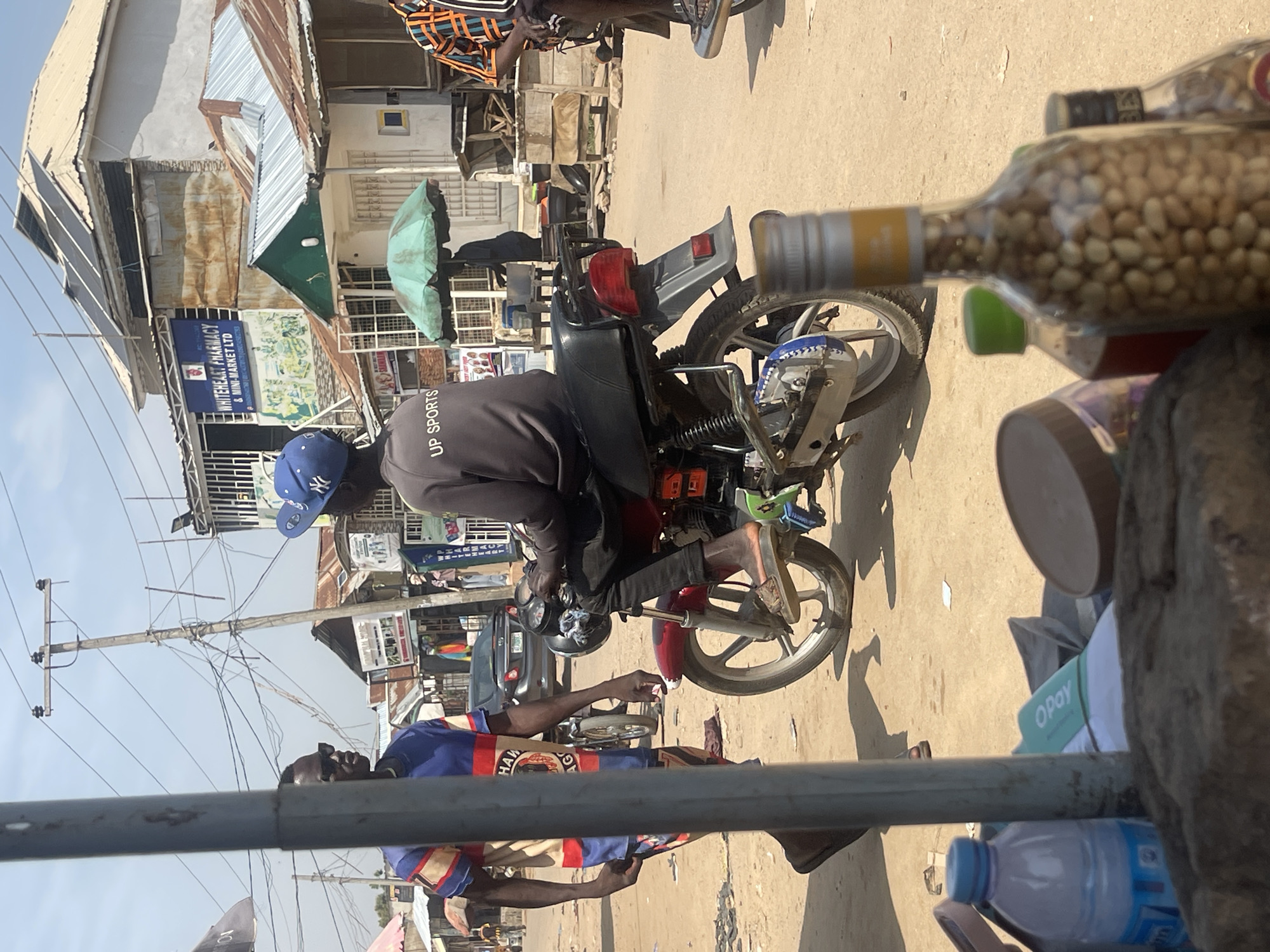 The image shows a street scene in Abuja, Nigeria, under a bright sky, suggesting a sunny day. The primary subjects are several individuals and a motorcycle. One person, wearing a blue baseball cap, is seated on the motorcycle, which is a common mode of transport in the area. Another person, in the foreground, is wearing a colorful striped shirt and appears to be looking down. In the background, there are various buildings, some with corrugated metal roofs, and market stalls, indicative of a bustling urban environment. Power lines crisscross the sky, a common sight in many Nigerian cities.
There is visible text on signs and merchandise. One sign reads "WHITEHEART PHARMACY & MINI-MARKET LTD," suggesting a commercial area. Another sign appears to say "UP SPORTS" on the back of a person's jacket. A bottle filled with nuts is prominently featured in the foreground, partially obscuring other objects. The overall impression is a candid snapshot of everyday life in a Nigerian city, with a focus on people, their mode of transportation, and the commercial activity. The scene appears to be during daylight hours.