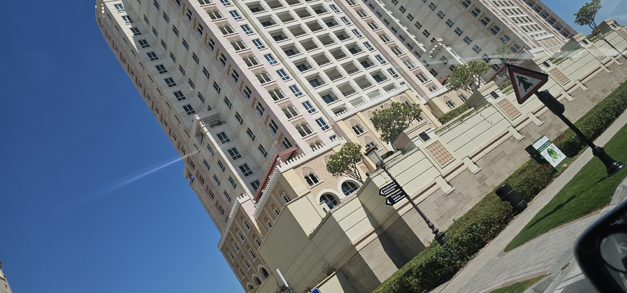 The image shows a low-angle, tilted view of a modern residential building in Doha, Qatar. The building is tall, with many floors and numerous windows. The facade is a light color, with decorative elements that suggest a contemporary architectural style. There is a clear blue sky with a faint light streak visible. In the foreground, there is a sidewalk, green grass, and some bushes. A street sign with Arabic text and directional arrows is visible, as well as a triangular traffic sign depicting a person in a traditional dress. The scene appears to be taken from a vehicle, as a portion of a car's interior is visible in the bottom left corner.