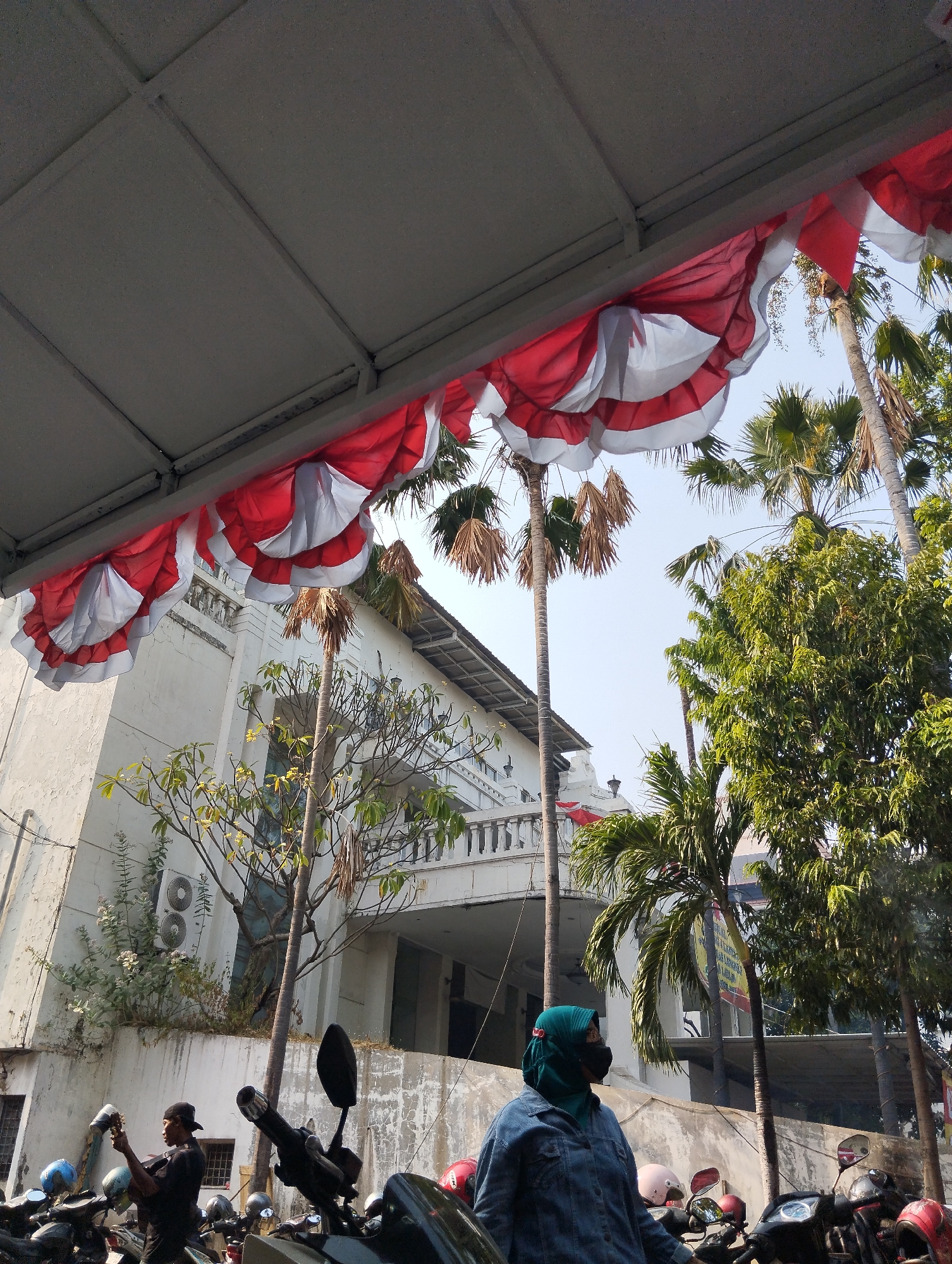 The image depicts a street scene in Surabaya, Indonesia, characterized by a building adorned with red and white pennants, possibly for a national celebration. The building, with its white facade and balconies, appears to be an older structure. Palm trees and other lush greenery surround it, suggesting a tropical environment. In the foreground, several motorcycles are parked, with two individuals visible. One person, wearing a dark t-shirt and a cap, is standing and holding an object. Another individual, wearing a teal hijab and a denim jacket, is standing next to a motorcycle. The sky is clear and blue, indicating a sunny day. The overall impression is of a vibrant urban setting, likely in a historical or culturally significant area of Surabaya.