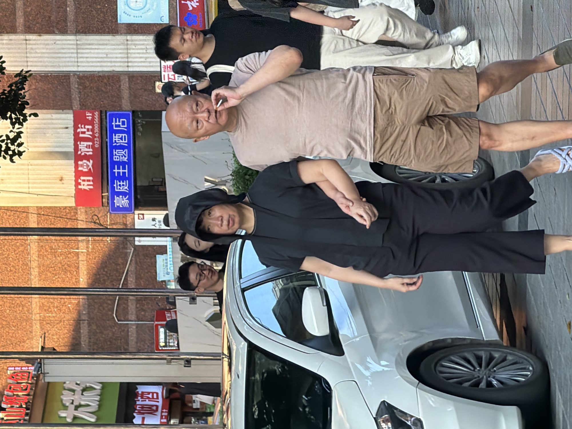 In a street scene in Yuzhong District, China, a group of people are gathered. A man in the foreground, with a shaved head and wearing a light brown t-shirt and beige shorts, is smoking a cigarette. He is standing next to a white car, and his arm is casually resting on the vehicle. Behind him, a person in a black hooded outfit is partially visible, looking towards the camera. Further back, another man with glasses is also looking in the direction of the camera. The background shows a building with signs, including one in red and white text that reads "柏曼酒店 4F" and a phone number, and another in blue that says "豪庭主题酒店". The overall atmosphere appears to be casual, with people standing around on a paved street. The lighting suggests it might be daytime, possibly late afternoon or early evening, given the shadows.