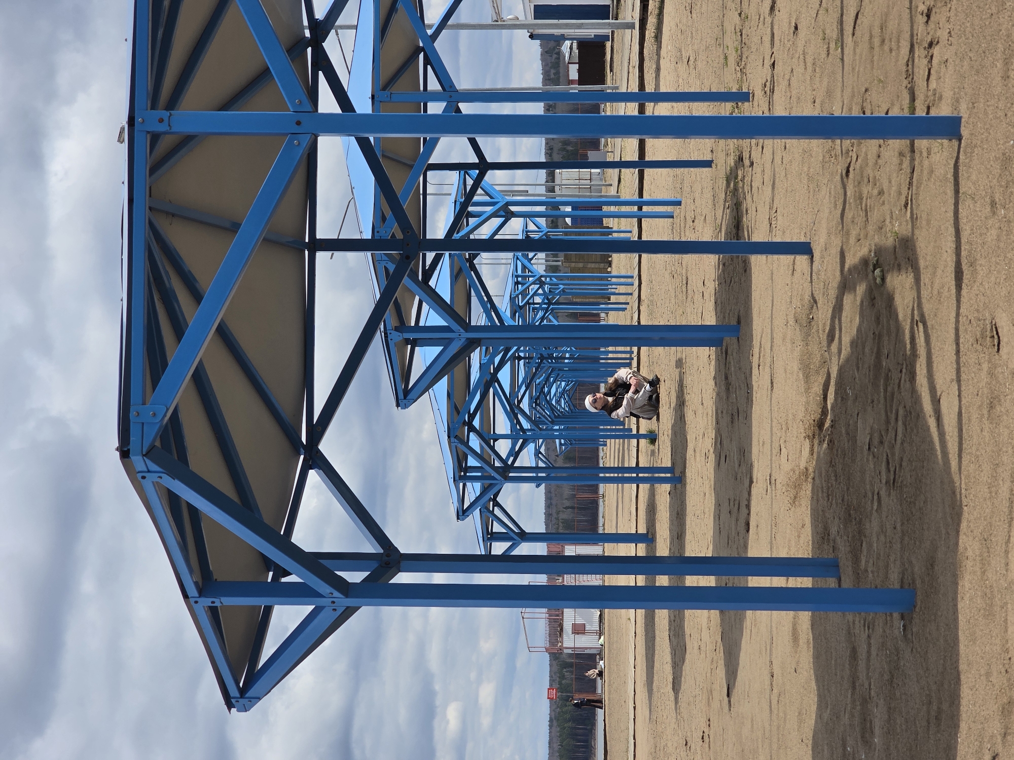 The image depicts a tranquil scene on a sandy beach or riverside area, likely in Chaikovsky, Russia. A long row of identical blue metal shade structures or canopies extends diagonally into the distance, providing a strong sense of perspective. Each structure consists of a robust blue frame supporting a light-colored, possibly beige or white, roof.
The ground is covered in light brown sand, showing subtle textures and ripples. Long, dark shadows are cast by the blue structures onto the sand, suggesting either early morning or late afternoon light. The sky above is partially cloudy, with patches of blue peeking through a soft gray overcast.
Two individuals are visible on the beach. In the mid-ground, a person wearing a light-colored jacket, pants, and a beanie is kneeling or squatting on the sand, appearing to be observing something on the ground. Further in the background, another person stands near the water's edge.
Beyond the beach, a body of water is visible, with a distant shoreline characterized by a strip of trees and several buildings, including a prominent white structure. The overall atmosphere is calm and quiet, with no obvious activities or events taking place. No discernible text is visible on any objects or signs within the frame.