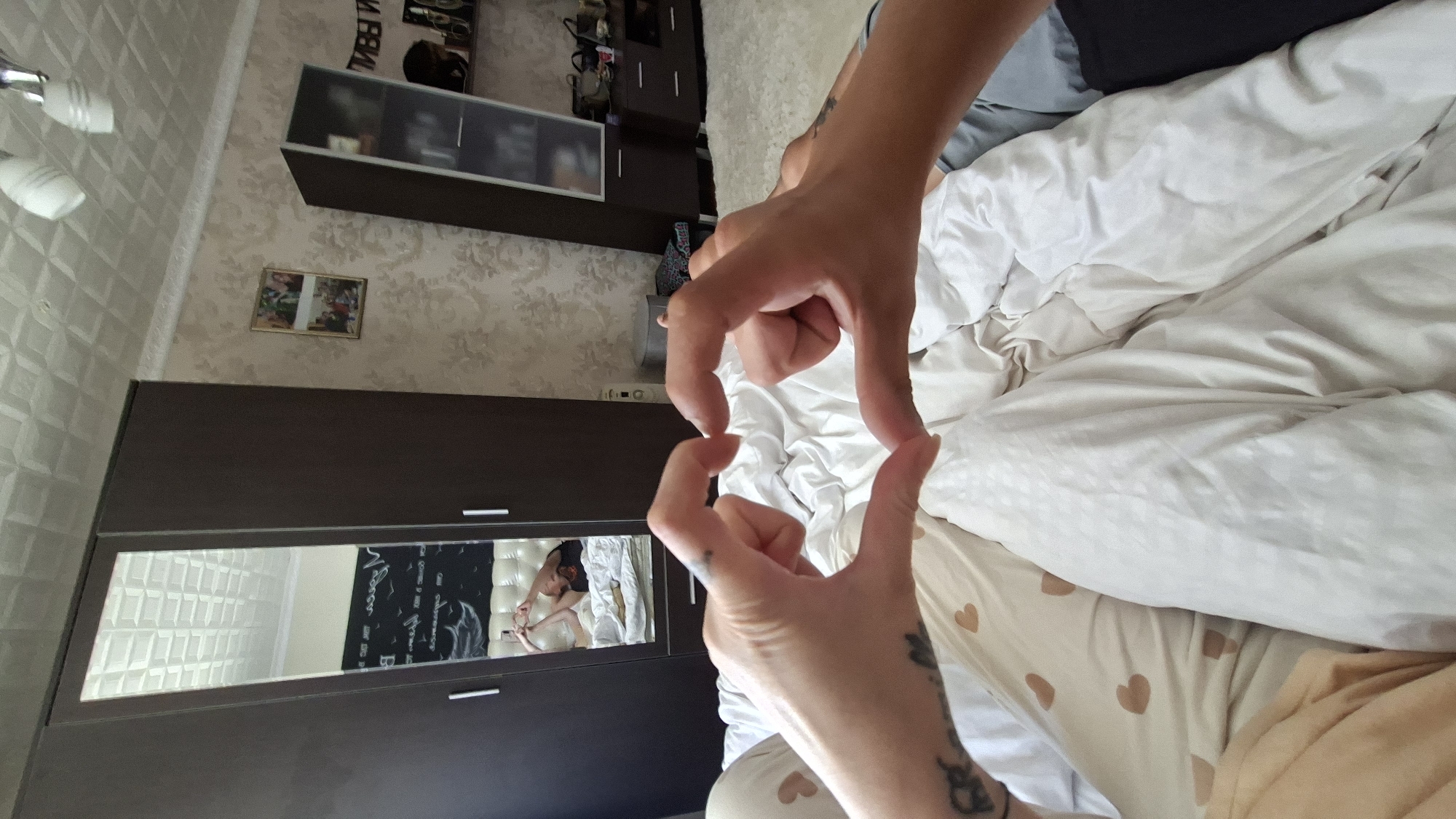 The image depicts a close-up perspective of two hands forming a heart shape over a bed covered with white and patterned bedding. In the background, a tall, dark brown wardrobe with a mirrored door is visible. Reflected in the mirror is a person lying on the bed, holding a phone and taking the photo. To the right of the wardrobe, there's a light-colored wallpaper with a floral pattern, and further to the right, a tall, dark glass-fronted cabinet containing various items. A ceiling light fixture with multiple bulbs is present in the upper part of the frame. The scene appears to be an indoor bedroom, possibly in a residential setting in Myadzyel, Belarus, based on the provided information.