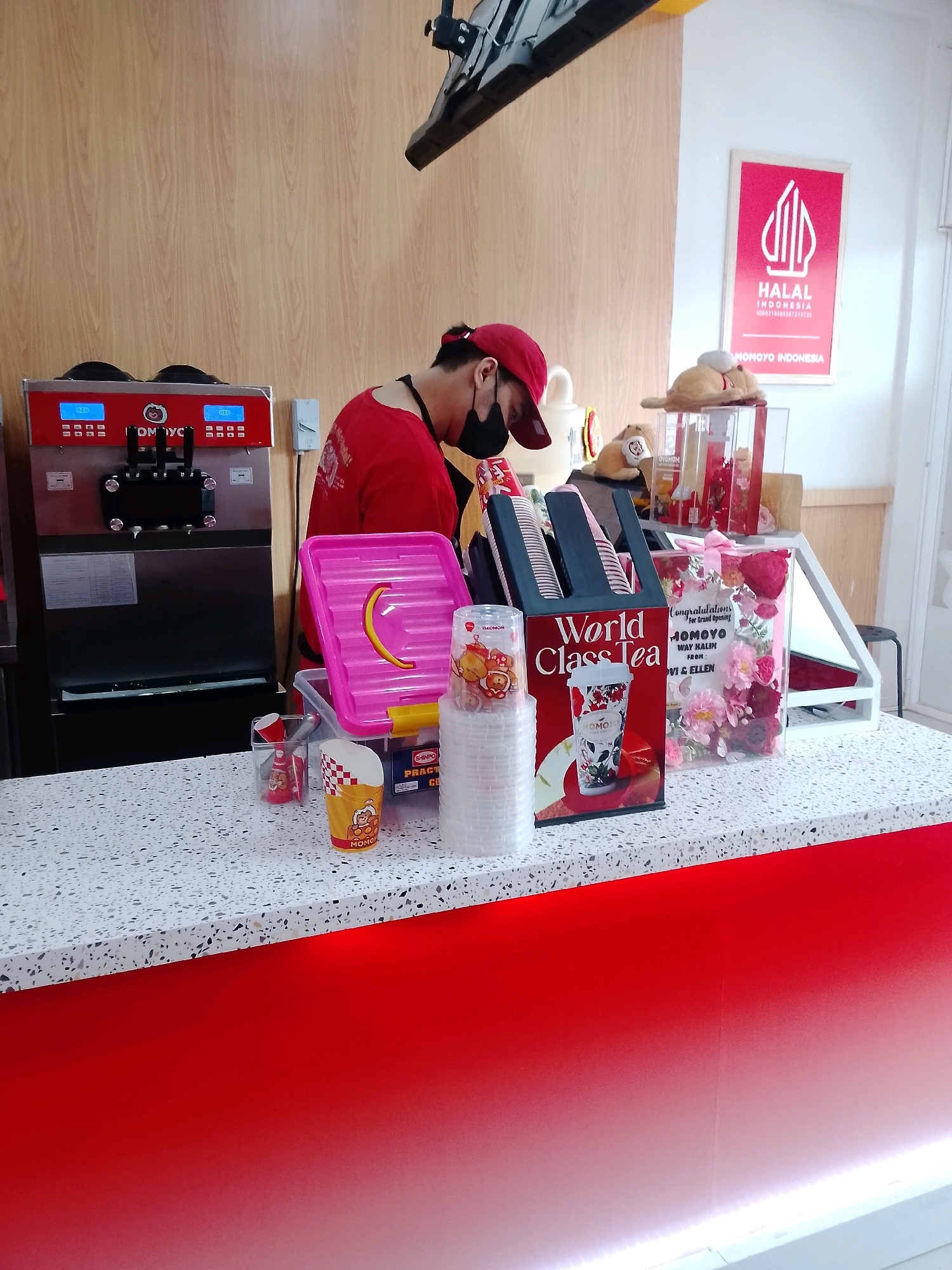 The image depicts the interior of a shop, likely a cafe or dessert store, with a focus on a counter area. A person wearing a red cap and a red shirt, with a black mask covering their lower face, is working behind the counter. They appear to be preparing or serving something, facing a soft-serve ice cream machine with two dispensers. The machine is branded with "OMOYO" and features digital displays.
On the counter, several items are visible:
* A pink tray with a yellow crescent shape.
* Stacks of disposable cups.
* A checkered cup with "MOMOYO" branding.
* A black holder with paper cups and a graphic of a tea cup labeled "World Class Tea".
* A clear box filled with artificial flowers, likely a gift, with a pink ribbon. Text on the box reads "Congratulations for Brand Opening MOMOYO", followed by names.
* Smaller decorative items and toys.
* A "HALAL INDONESIA" sign is mounted on the wall behind the counter.
The environment appears clean and well-lit. The counter has a speckled white surface, and the lower part of the counter is a vibrant red, with a gradient effect towards white at the bottom. The wall behind the counter is made of light-colored wood panels.
Based on the presence of a "HALAL INDONESIA" sign and the brand name "MOMOYO", which is known for its ice cream and beverage offerings in Indonesia, this scene is consistent with a Momoyo outlet. While the specific location within Bandar Lampung is not explicitly stated in the image, such an establishment would typically be found in commercial areas or shopping centers within the city.