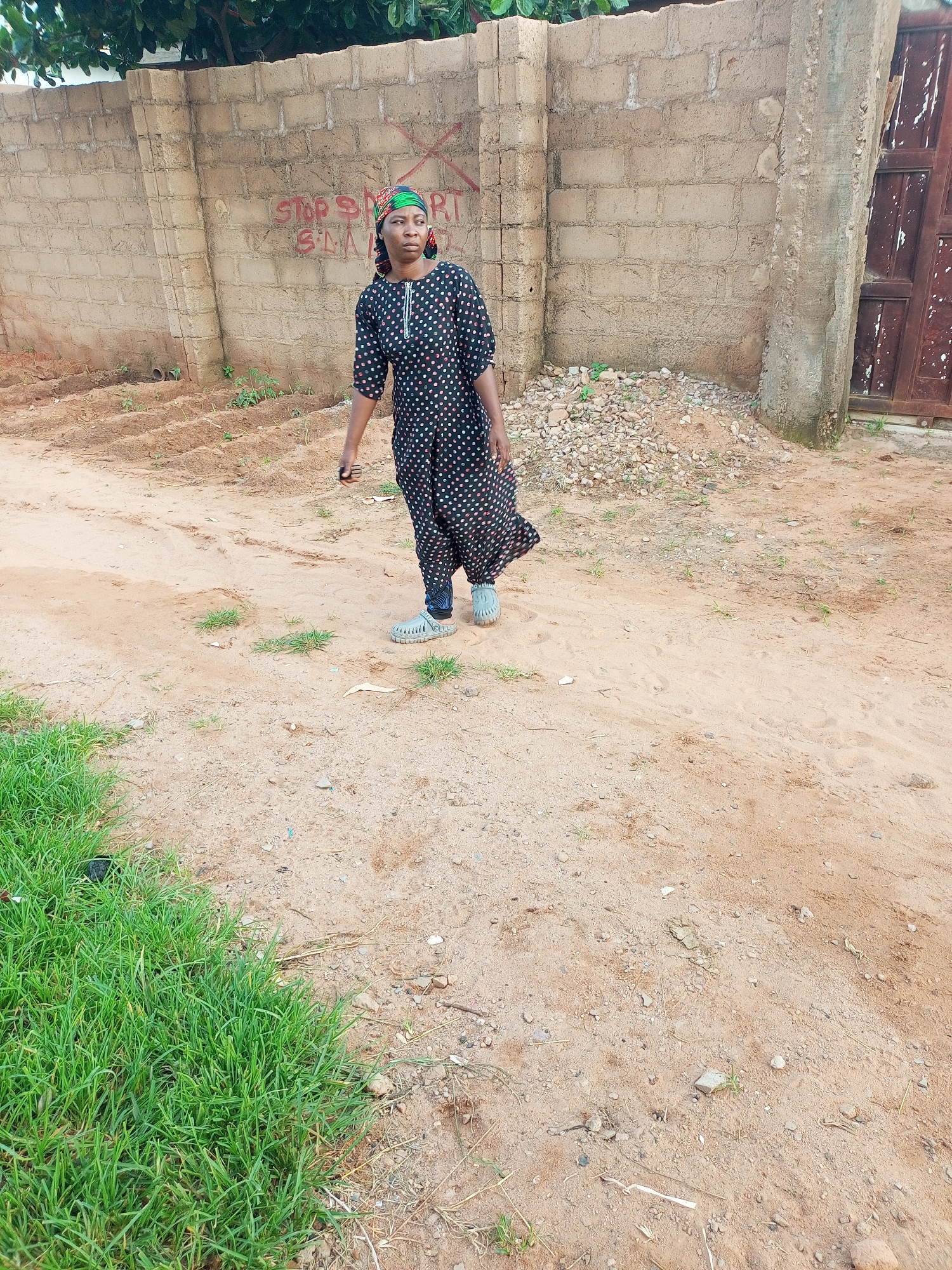 A Black female wearing a black and white polka dot dress and a green and red headscarf walks in front of a brick wall with graffiti. She is wearing light gray slip-on shoes. The wall is weathered and has a wooden door on the right side. The ground in front of the wall is dirt and sand with some sparse green grass and small stones. The background shows more trees and foliage. The scene appears to be outdoors, possibly in a rural or semi-urban area of Gombe, Nigeria.