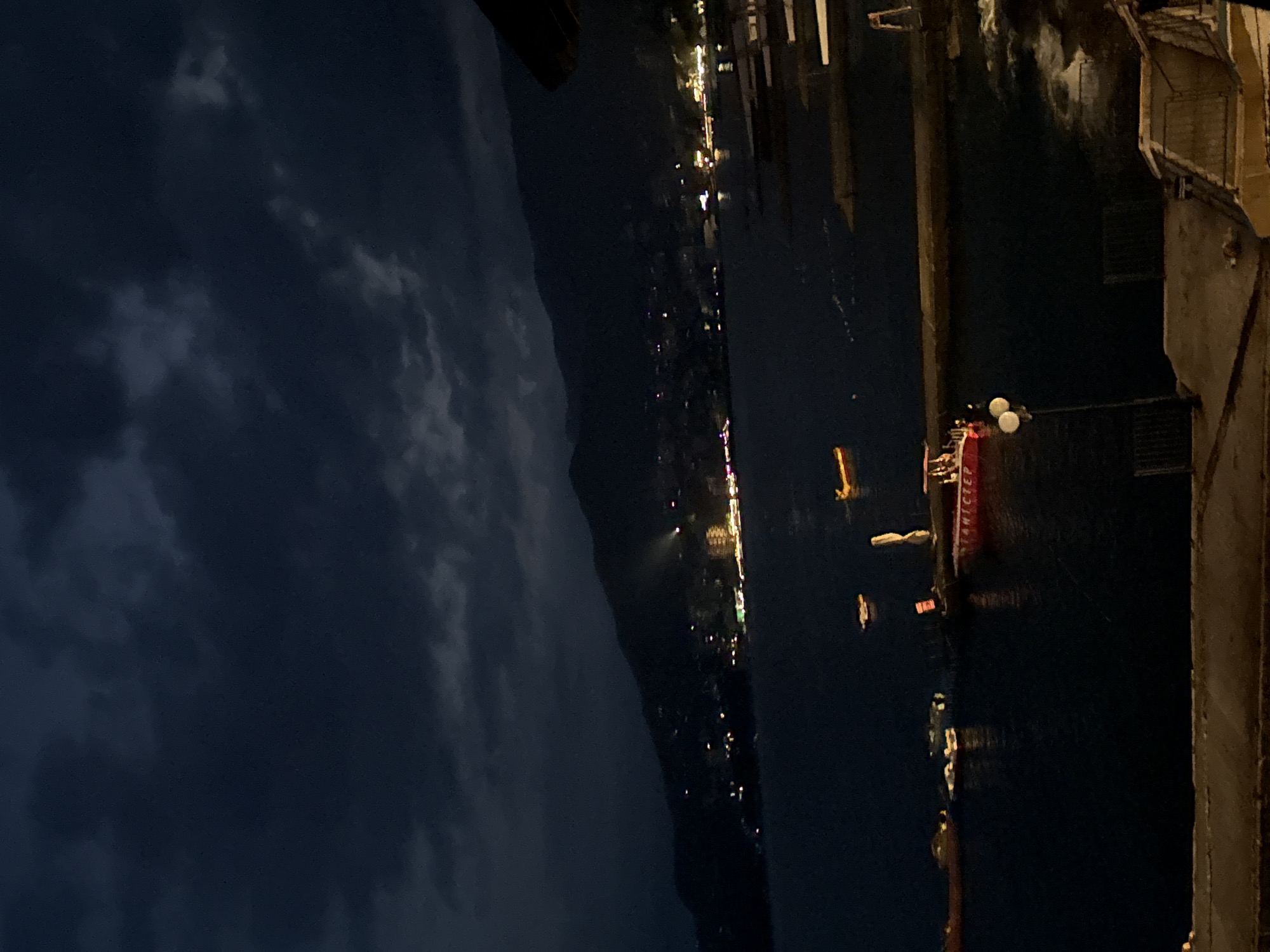 A nighttime, low-angle, medium shot captures a dark, cloudy sky over a body of water, possibly a bay or sea, with a dimly lit mountainous coastline in the background. A few boats are visible on the water, including a small red vessel with lights. The coastline is dotted with lights from buildings, and a spotlight shines from the mountainside. In the foreground, a concrete structure with a railing and a lamp post with two globes are partially visible, suggesting a waterfront promenade or pier. The overall atmosphere is dark and somewhat moody, illuminated by scattered artificial lights. The location is identified as Yalta, Ukraine, a coastal city known for its mountainous landscape and harbor.