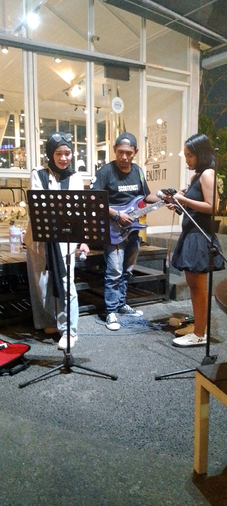 In Semarang, Indonesia, a street performance is underway with three individuals in front of a modern building with large glass windows. On the left, a woman wearing a black hijab and sunglasses stands behind a black music stand, holding a microphone in her right hand. To her right, a man in a dark t-shirt with the word "SCOOTERIST" printed on it, and a backward baseball cap, plays a purple electric guitar. In front of him, a blue cable lies coiled on the ground. On the far right, a young woman in a black sleeveless dress and white sneakers holds a microphone. The background shows a seating area with wooden tables and chairs, and the building appears to be a cafe or restaurant, with text on the glass that reads "THIS IS OUR SECOND HOME WE SPEND ENJOY IT." The scene takes place at dusk or night, with artificial lighting illuminating the area.