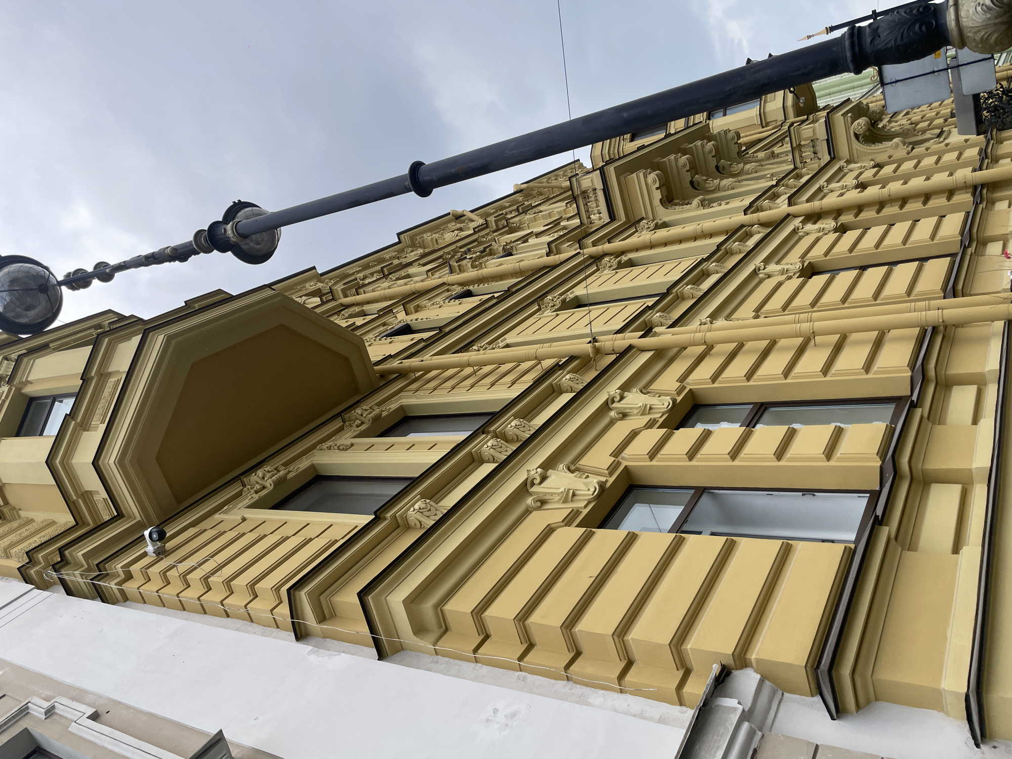 The image presents an upward-angled view of a classical architectural building facade in Saint Petersburg, Russia, under an overcast, cloudy sky. The building is predominantly a warm yellow or ochre color, richly detailed with ornate moldings, cornices, and decorative corbels above and below its many rectangular windows. The window frames are dark, providing a stark contrast to the light-colored facade, and the windows themselves show reflections or darkness within.
A prominent, dark-colored street light structure spans horizontally across the upper part of the frame, featuring two large, spherical light fixtures and several smaller, decorative elements along its length. Various pipes and conduits, including horizontal segments across the facade and a thicker vertical black pipe, are visible. On one of the lower ledges, a small white security camera is mounted, with a thin wire running past it. The bottom section of the image reveals a lighter, possibly white or light grey, lower part of the building or an adjacent structure.
No people are visible, and no discernible text is present. The daytime lighting is soft and diffused due to the overcast conditions. The scene focuses on the intricate architectural details and the imposing scale of the building.