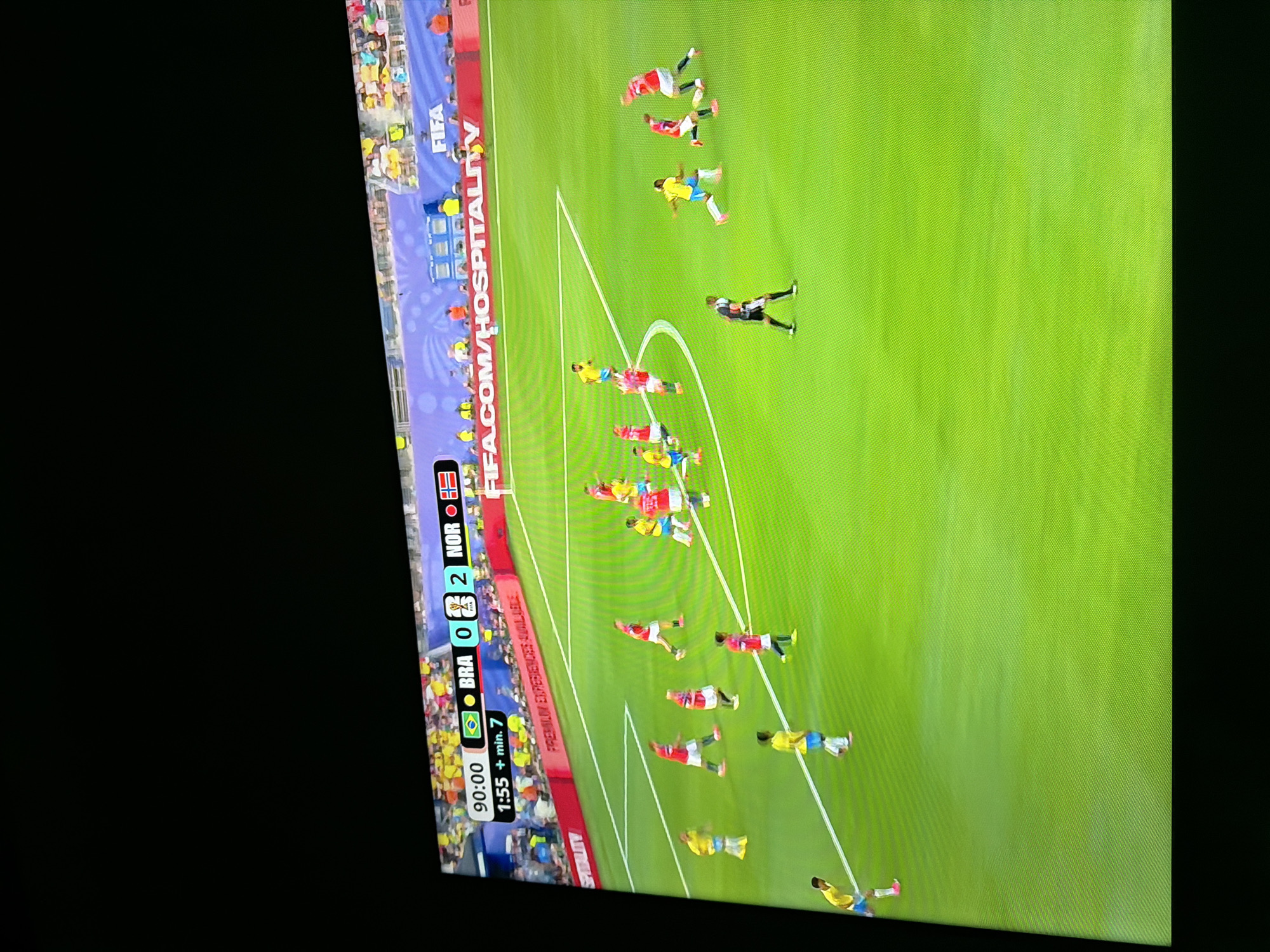 The image displays a broadcast of a soccer match. The scoreboard at the top indicates the game is at 90:00 plus 7 minutes of stoppage time. The score is BRA 0, NOR 2. The field is green with white lines marking the boundaries and penalty areas. Several players in yellow and red uniforms are visible on the field, engaged in the game. Spectators are present in the stands behind the field, and banners with the FIFA logo and "FIFA.COM/HOSPITALITY" are displayed. The image is captured from a screen, suggesting it is a photograph of a television or monitor displaying the match. There is no information in the image to suggest any connection to the city of Ognianovo, Bulgaria.