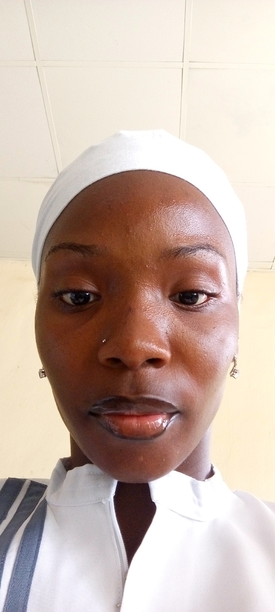 A close-up, high-angle shot shows a person with dark skin, wearing a white head covering and white clothing with a striped detail on the shoulder. The person has dark eyes, a nose piercing, and dark, glossy lips. The background is a light yellow wall and a white ceiling with grid lines. The location is Unknown, Unknown.