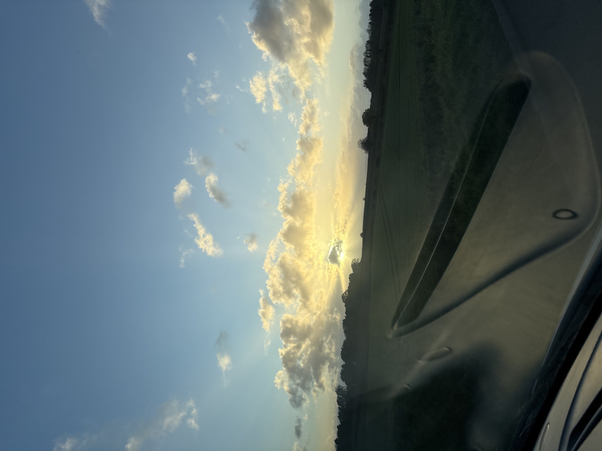 The image, taken from inside a vehicle, shows a view of the sky and landscape near Ruyton-XI-Towns, United Kingdom. The sky is predominantly blue with scattered, fluffy white clouds that are illuminated by the setting sun, which is partially obscured by the clouds near the horizon. Sun rays emanate from behind the clouds. Adjacent to the sky, on the right side of the image, is a field or agricultural area with a line of trees visible along the horizon. The interior of the vehicle, specifically part of the dashboard and possibly the windshield, is visible in the lower right corner, casting a reflection and obstructing some of the view. No people are directly visible. The scene depicts a natural environment at sunset.