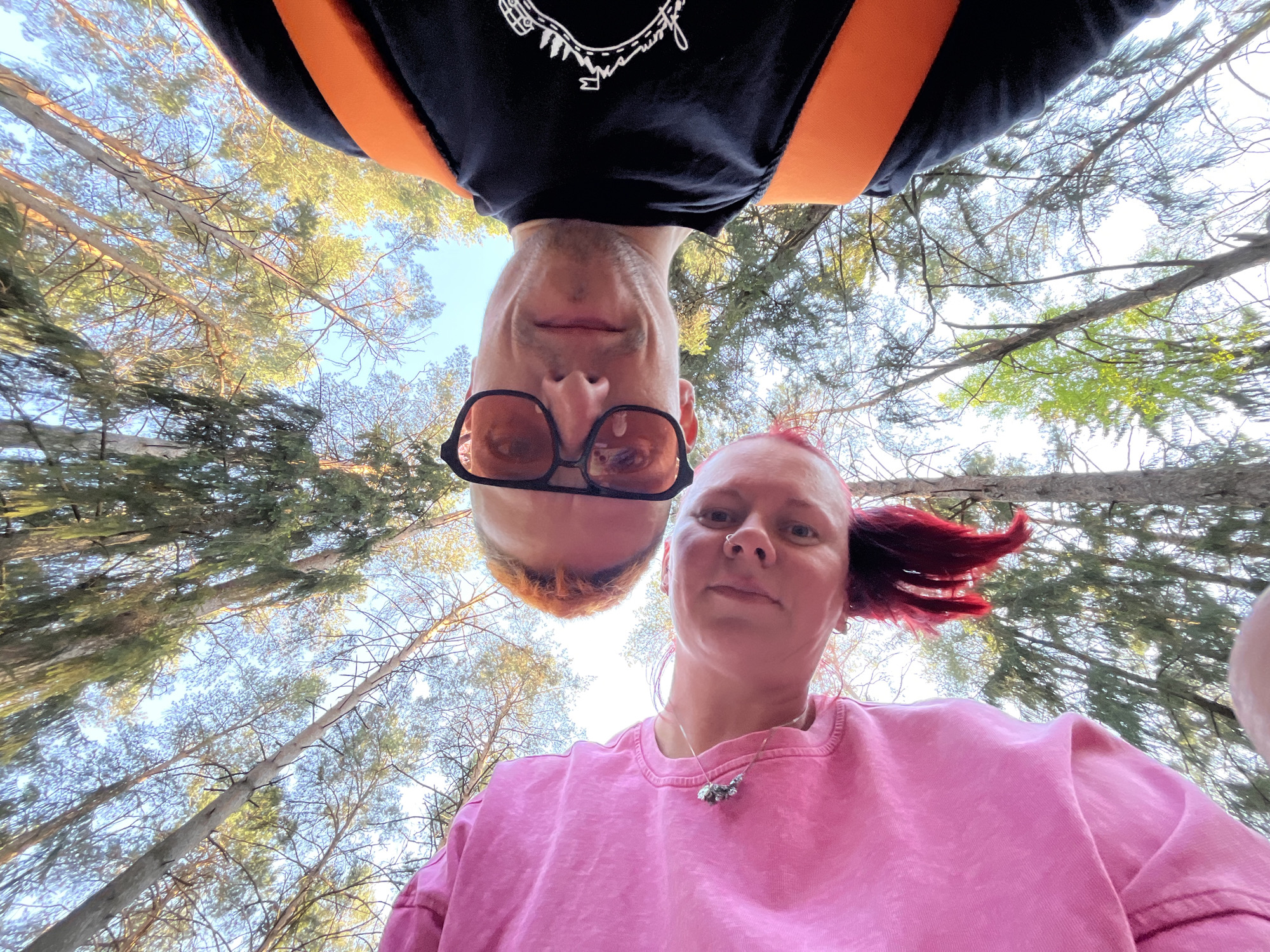 From a low angle, two people are photographed, appearing upside down, in an outdoor forest setting. A person with short, light-colored hair and wearing dark-rimmed glasses is positioned towards the top of the frame. The second person is wearing a pink top. Visible behind them is a dense forest canopy with a sky visible through the leaves. The location is likely in Chaikovsky, Russia.