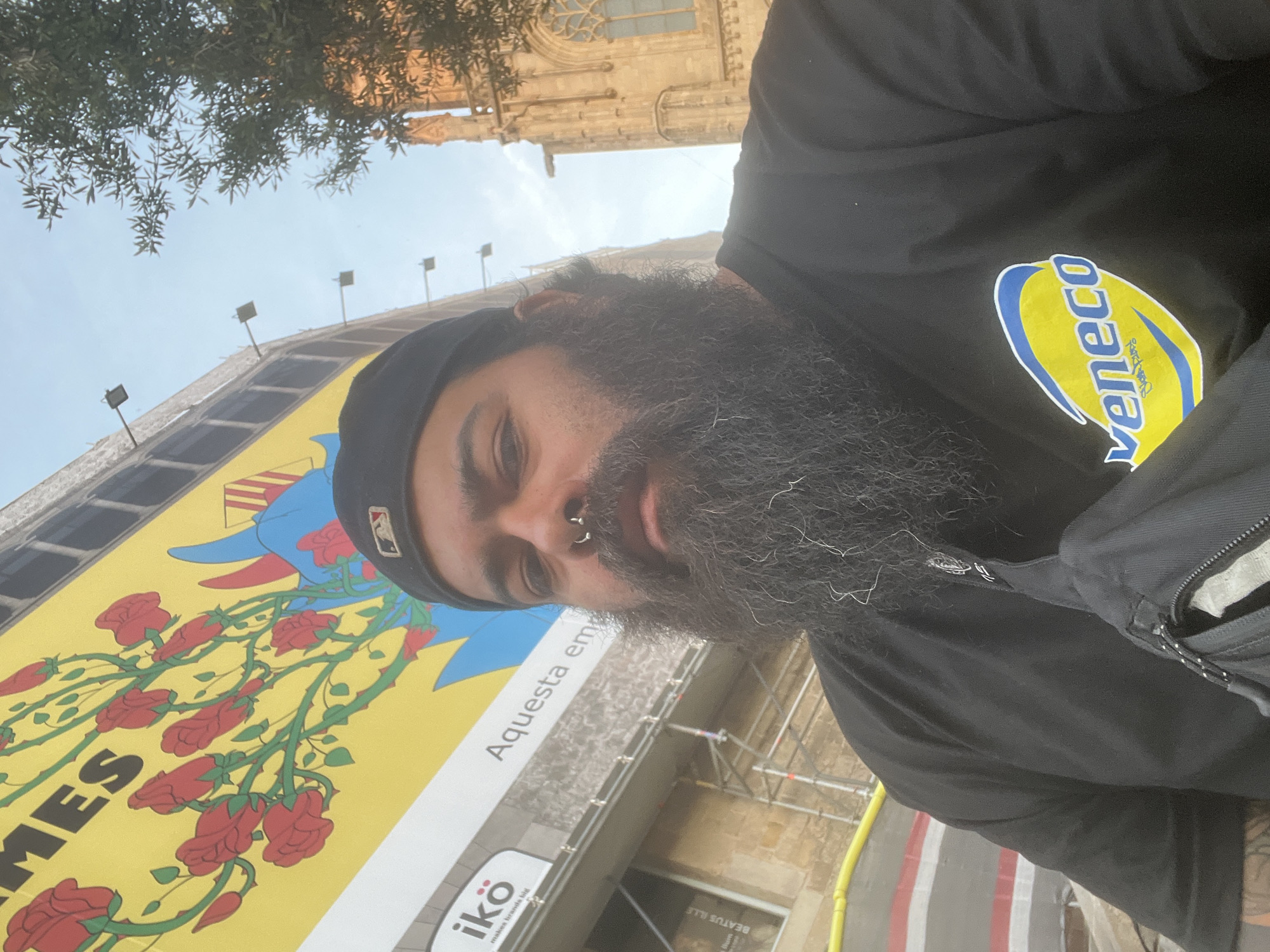 A man with a full beard and a nose ring is taking a selfie in Barcelona, Spain. He is wearing a black shirt with the "Veneco" logo and a black baseball cap with the MLB logo. Behind him is a large yellow building facade adorned with red roses and a blue figure, possibly related to a promotional campaign with the word "mes" visible. The building is under some form of construction. Part of the Barcelona Cathedral is visible in the top right corner of the image. The sky is overcast. Scaffolding is present.