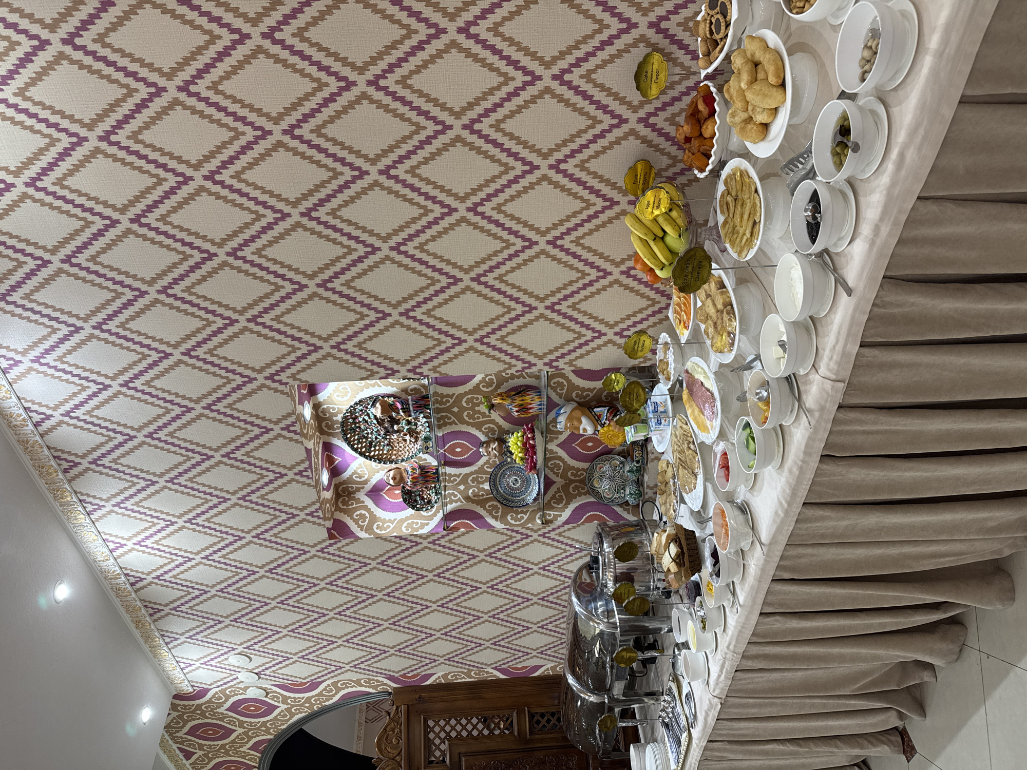 The image depicts a lavish breakfast buffet in what appears to be a hotel or banquet hall in Samarkand City, Uzbekistan. The ceiling is adorned with a geometric diamond pattern in beige and purple. The buffet table is laden with a variety of food items, including fresh fruit (bananas, citrus), pastries, nuts, olives, and various other dishes served in white bowls and platters. Beverage dispensers and stacks of cups are visible at one end of the table. The table is covered with a light beige tablecloth that drapes to the floor. The room features ornate woodwork along the lower wall, and a decorative lighting fixture hangs from the ceiling. The overall impression is one of abundance and hospitality.