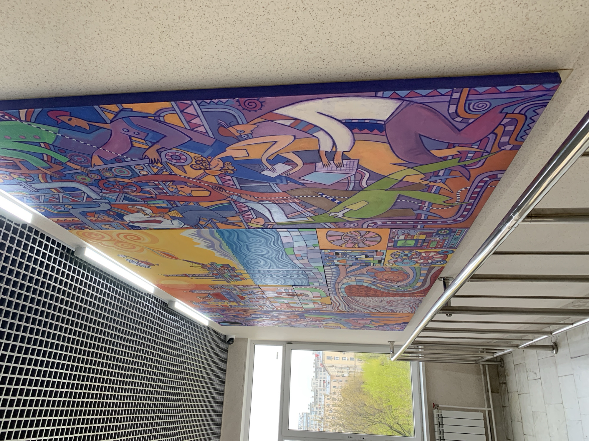 The image displays a vibrant, abstract mural covering a ceiling, seen from an angled, upward perspective, within what appears to be an indoor stairwell or corridor. The mural features stylized human figures, intricate mechanical elements like gears and pipes, and organic forms such as bird-like creatures and plant-like shapes. One figure, wearing light clothing, is hunched over a laptop, appearing to work or study, with papers scattered nearby. Another figure in darker attire is integrated with industrial-looking machinery. The mural transitions into landscapes, including blue wavy patterns representing water and yellow/orange areas suggesting land, interspersed with geometric patterns that might depict urban structures or technological grids. The overall aesthetic is characterized by bold outlines and a rich palette of purples, blues, oranges, greens, and yellows, suggesting themes of human endeavor, technology, and nature.
To the left, a dark, grid-patterned wall runs along the base of the mural, illuminated by a long, bright LED light fixture. On the right, a metallic railing structure, likely part of a staircase, extends from the floor upwards. A large window occupies the lower right, revealing an outdoor scene. Outside, green trees with fresh foliage indicate spring or early summer, and tall, multi-story urban buildings are visible under a bright, possibly overcast, daytime sky. Below the window, a modern white radiator is visible. A small white security camera is mounted high near the window frame. The scene is consistent with an indoor public or residential space in Moscow, Russia. There is no visible text.