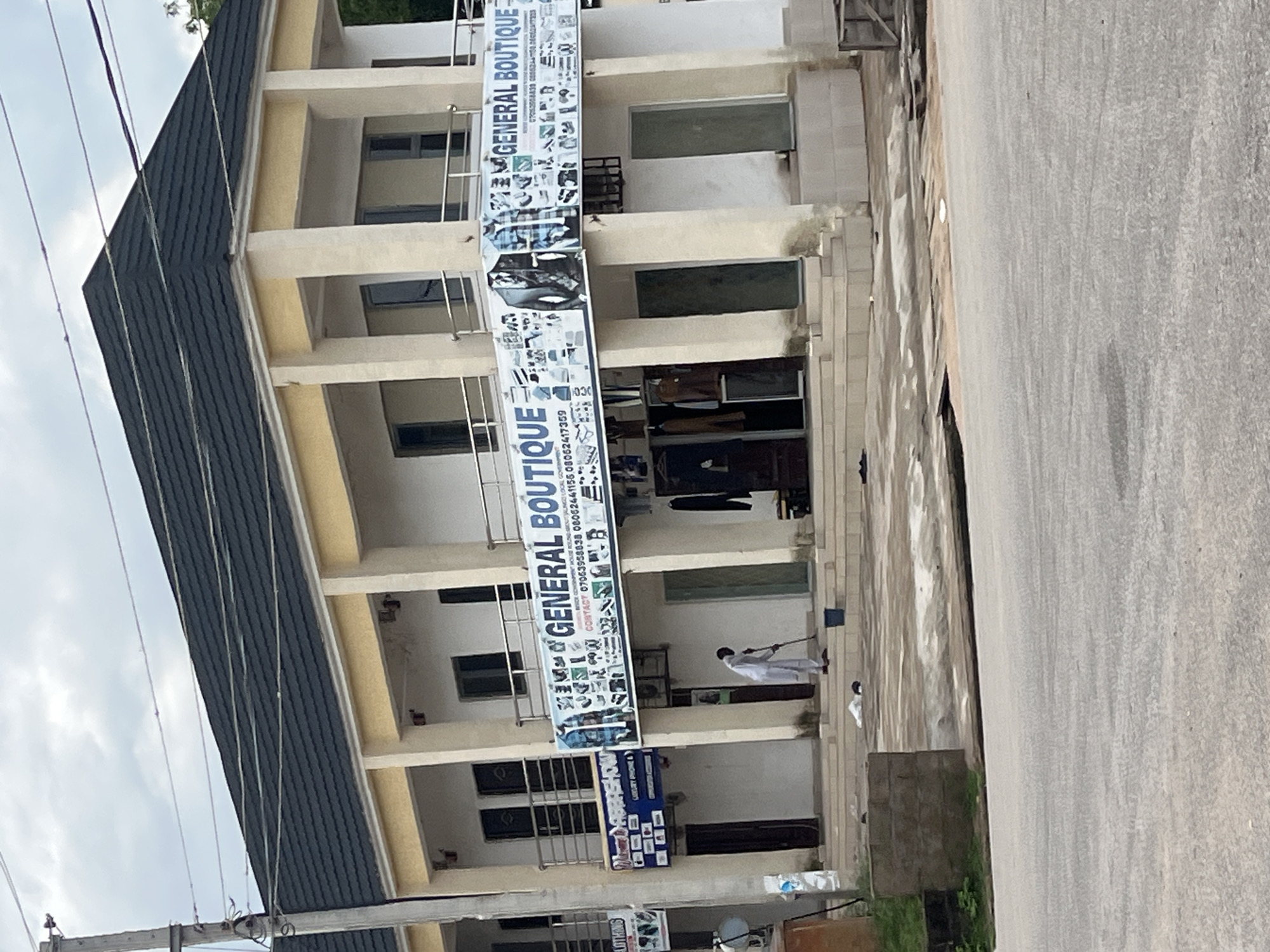 The image shows a multi-story building with a dark tiled roof, painted in light yellow and white. A banner across the second floor reads "GENERAL BOUTIQUE" and displays various clothing items and contact numbers. On the ground floor, a person dressed in white is sweeping the tiled entryway with a broom and a blue bucket. The building appears to be a commercial establishment, likely a clothing store, situated on a paved street in Jalingo, Nigeria. The scene is captured in daylight, under an overcast sky, with some greenery visible on the left side of the frame.