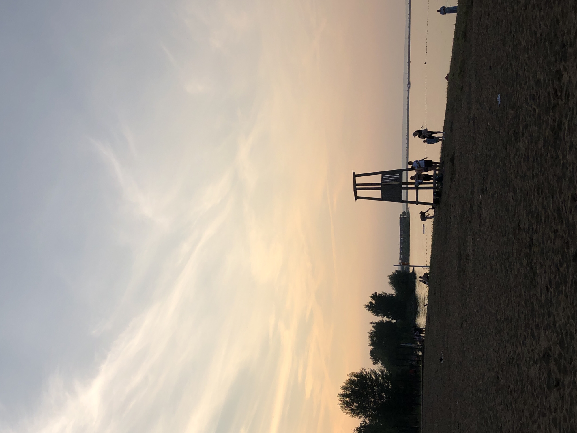 This is a vertical, low-angle, close-up shot of a beach at sunset. The sky is a blend of pale blue and soft peach, with wispy clouds stretched across it. In the foreground, the sandy beach is dark and textured, with a few scattered pieces of debris. A lifeguard stand, silhouetted against the sky, stands on the left side of the frame.
Further into the scene, a group of people are gathered on the beach and in the shallow water. Some are standing and talking, while others are partially submerged. The water is calm and reflects the warm hues of the sunset. Trees line the edge of the beach, providing a dark silhouette against the horizon. In the distance, buildings and more trees are visible across the water, suggesting a town or city on the opposite shore.
The overall mood is peaceful and serene, with the warm colors of the sunset contributing to a sense of calm. The presence of people enjoying the beach indicates a recreational setting, likely a public beach. The time of day is clearly sunset, with the low angle of the sun casting long shadows and bathing the scene in warm light. There is no visible text in the image.