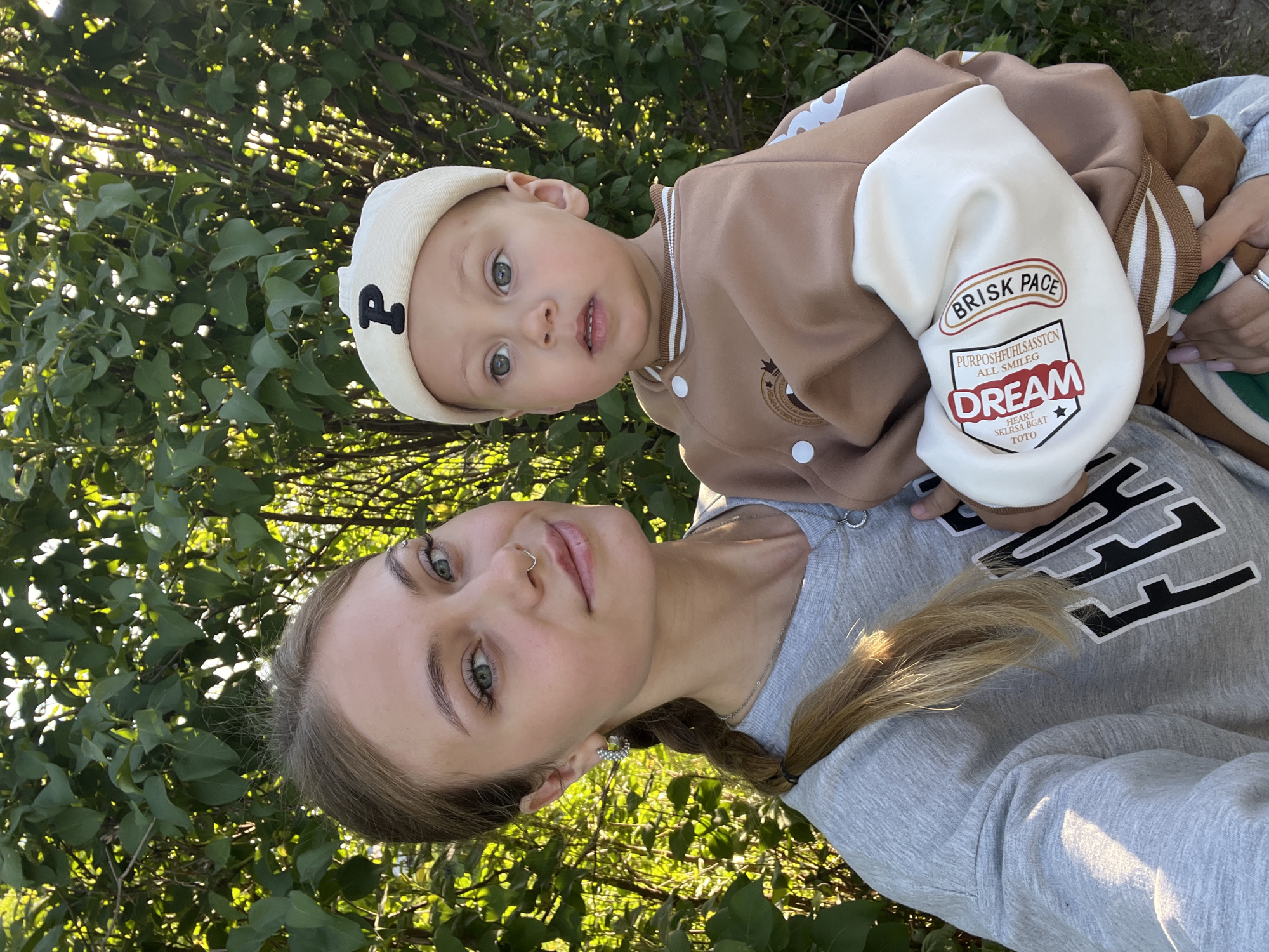 A young woman with light brown hair braided and a silver nose ring is holding a young child with blonde hair and a white hat. The child is wearing a brown and white jacket with the words "BRISK PACE" and "DREAM" on it. The woman is wearing a grey sweatshirt. They are outside in front of green foliage. The location is Фоки, Russia.