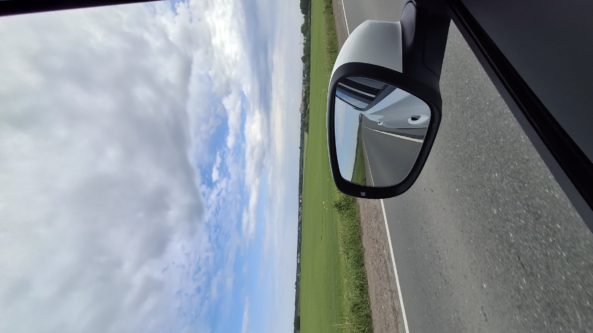 This image captures a wide-angle view from inside a moving vehicle, looking out the passenger side window during daytime in Pigelevo, Russia.
The primary subject is the car's white side mirror, which occupies the mid-right portion of the frame. The mirror's dark trim frames a reflection of the asphalt road receding behind the car, along with a partial view of the car's white body. To the immediate right of the mirror, a dark section of the car's interior or door frame is visible.
Beyond the car, the landscape is dominated by a vast, bright green field stretching horizontally across the middle of the scene, extending towards a distant horizon line where faint trees and structures can be discerned. The field borders a grey asphalt road, which features a clear white line marking its shoulder on the right.
The sky occupies the upper half of the image, presenting a mix of bright blue patches and scattered, fluffy white and light grey clouds, indicating partly cloudy weather. No people or visible text are present in the image. The scene conveys a sense of serene travel through an open, rural landscape.